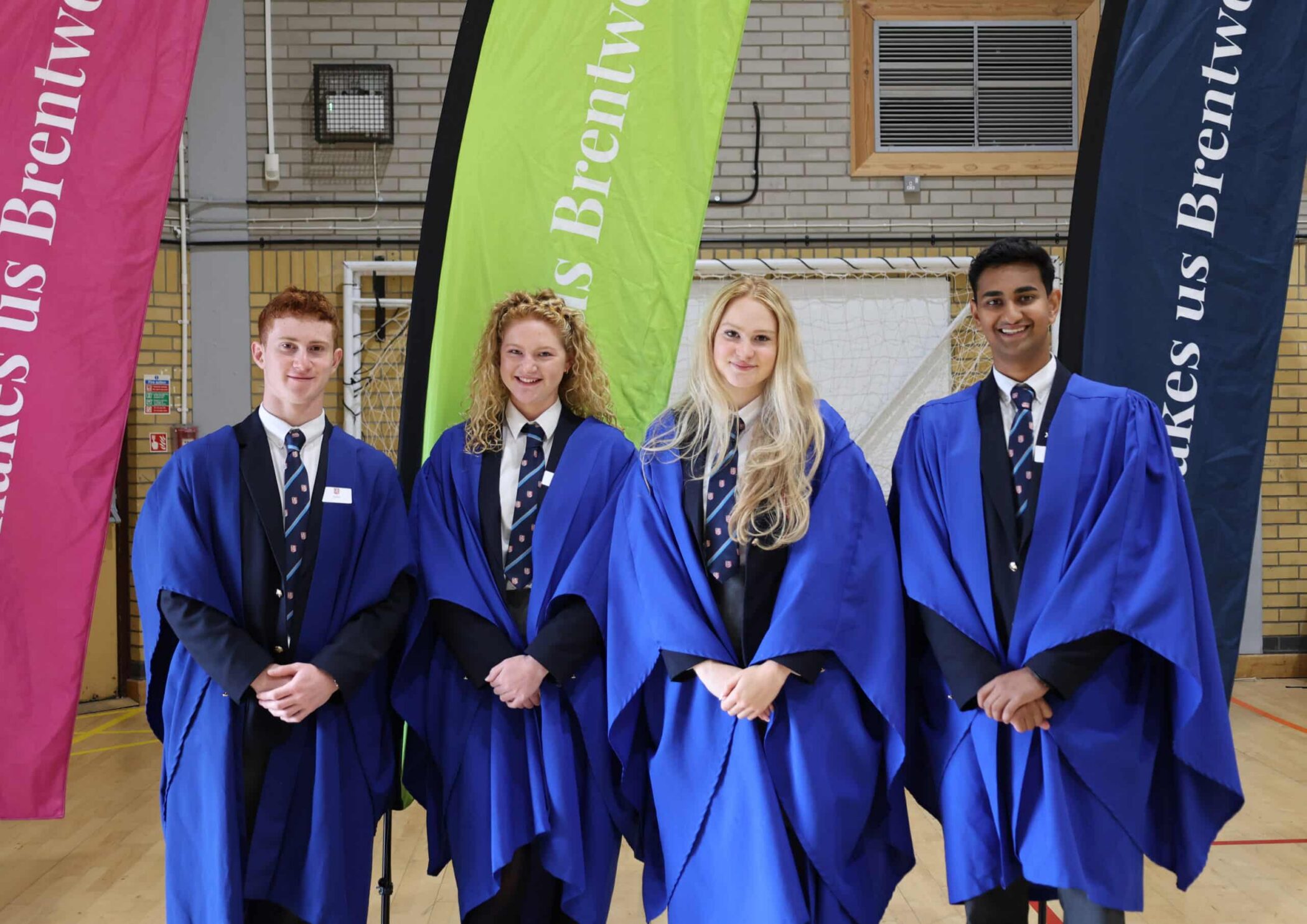Four students in blue graduation gowns stand indoors, smiling, in front of colourful “Brentwood” banners. Dressed in striped ties and formal attire, they pose at Open Events in a sports hall-type setting.