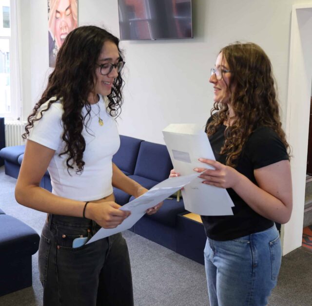 Two young women with long curly hair and glasses stand indoors, smiling at each other whilst holding papers. They appear to be sharing good news in a relaxed lounge area with blue sofas.