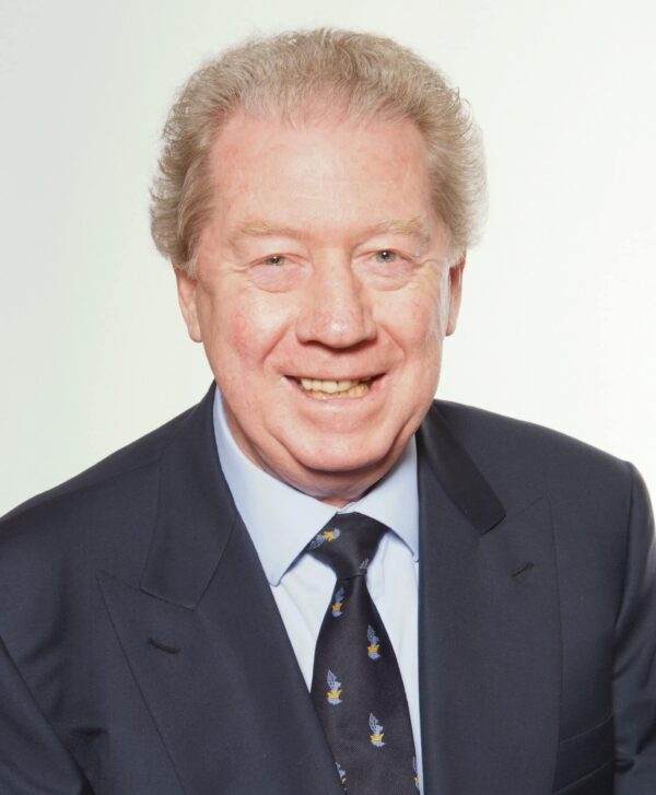 A smiling older man with light skin and short, curly grey hair wearing a dark suit, light blue shirt, and a patterned tie, posing against a plain light background.
