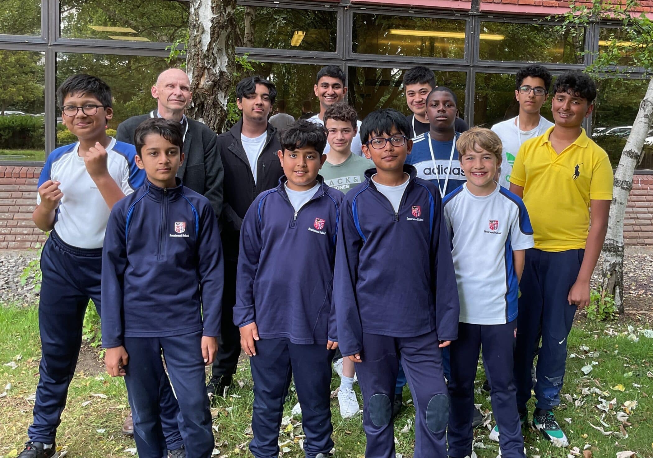 A group of teenage boys and one adult stand outside in front of a building, posing for a group photo. Most of the boys wear matching navy sports kits; some are in white shirts. They are smiling on a grassy area with trees.