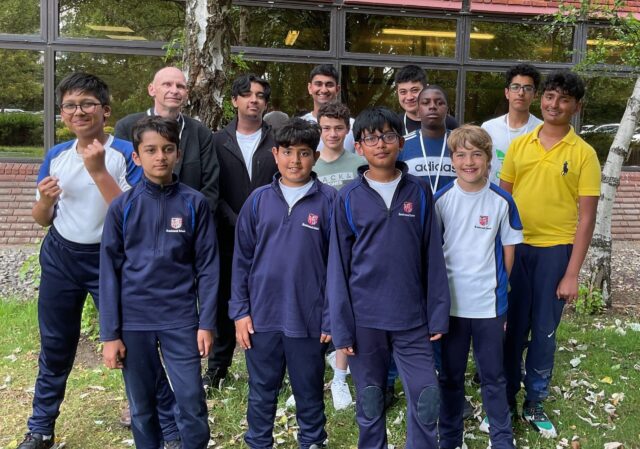 A group of teenage boys and one adult stand outside in front of a building, posing for a group photo. Most of the boys wear matching navy sports kits; some are in white shirts. They are smiling on a grassy area with trees.