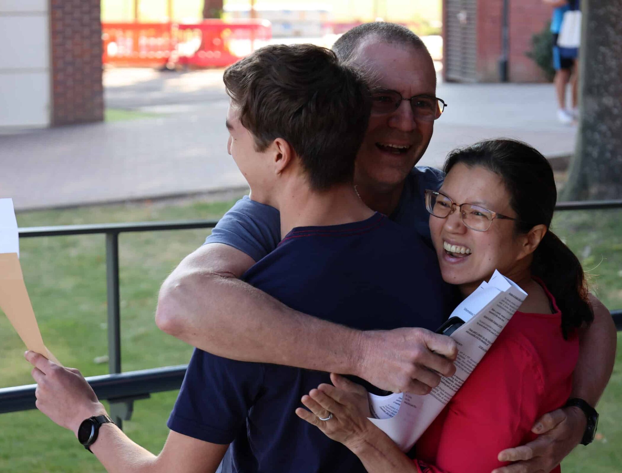 Three people are standing outdoors, smiling and sharing a joyful group hug. One person is holding papers, and all appear happy and celebratory. Trees and a building are visible in the background.