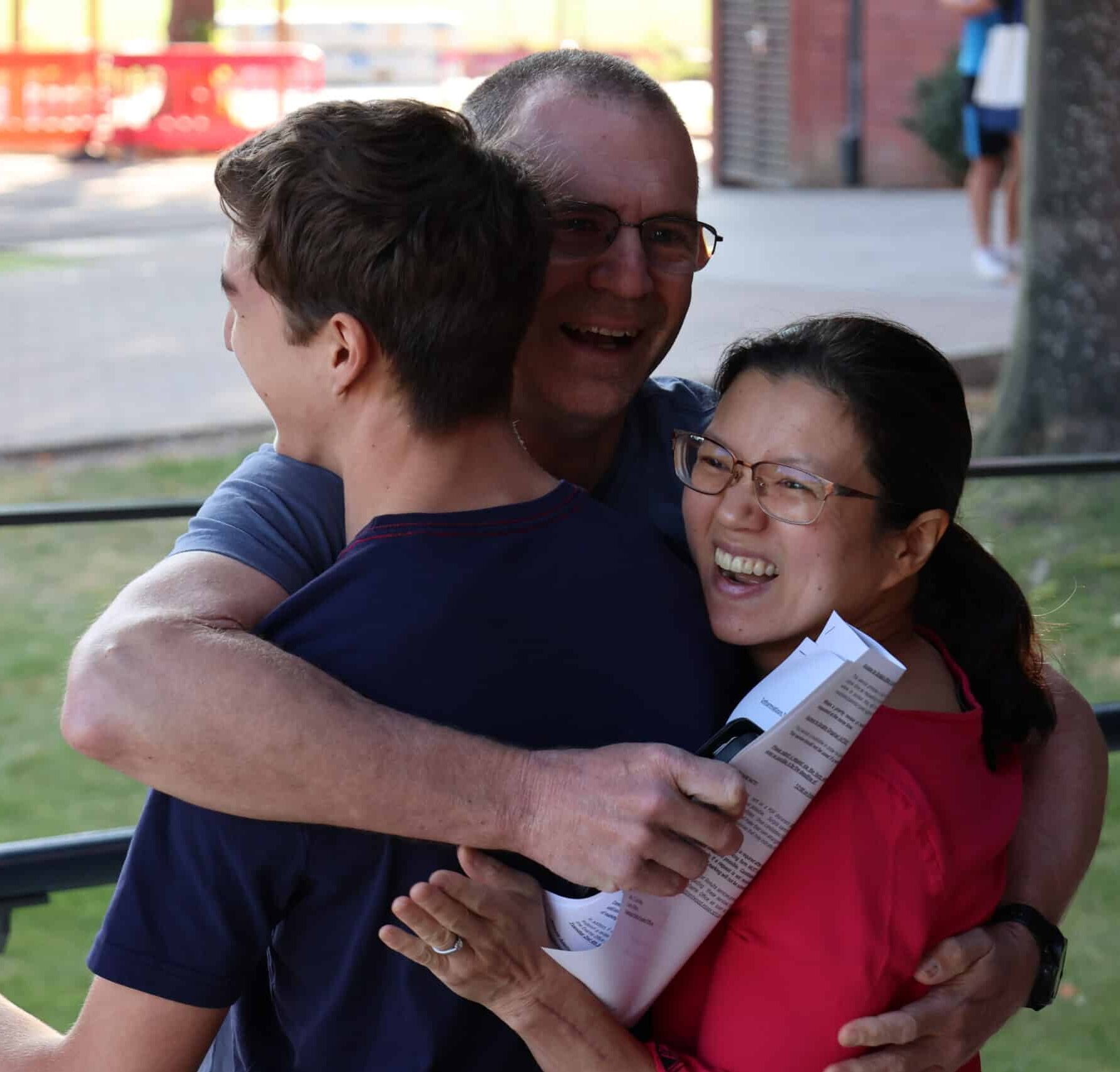 Three people are standing outdoors, smiling and sharing a joyful group hug. One person is holding papers, and all appear happy and celebratory. Trees and a building are visible in the background.