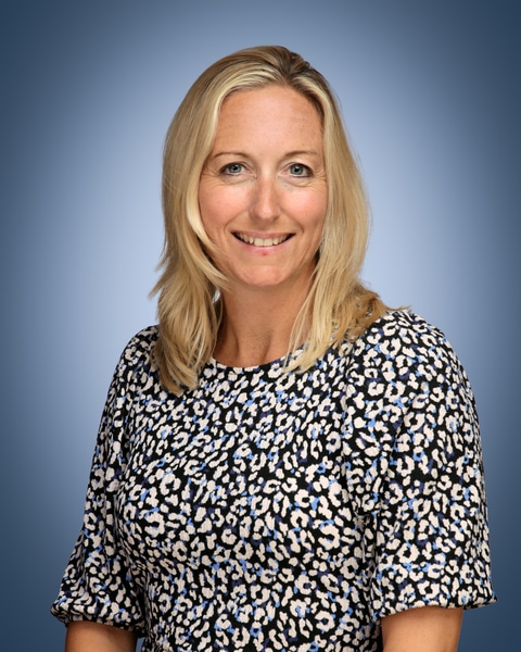 A woman with blonde hair, Assistant House Manager, smiles at the camera in a blue, white, and black patterned top against a blue gradient background.