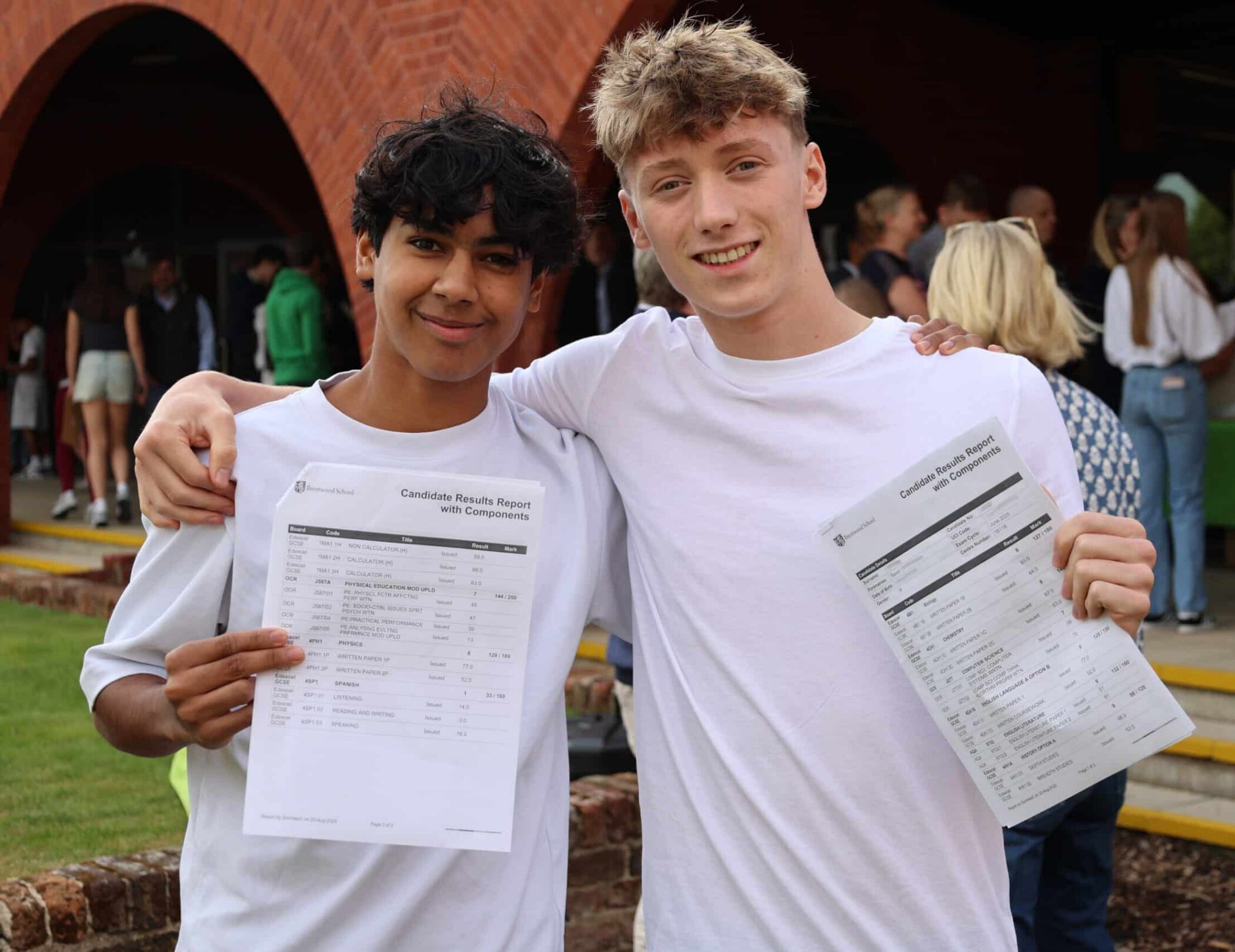 Two smiling teenagers stand arm-in-arm outdoors, proudly holding up their GCSE exam results papers. Other people are gathered in the background near a brick building.