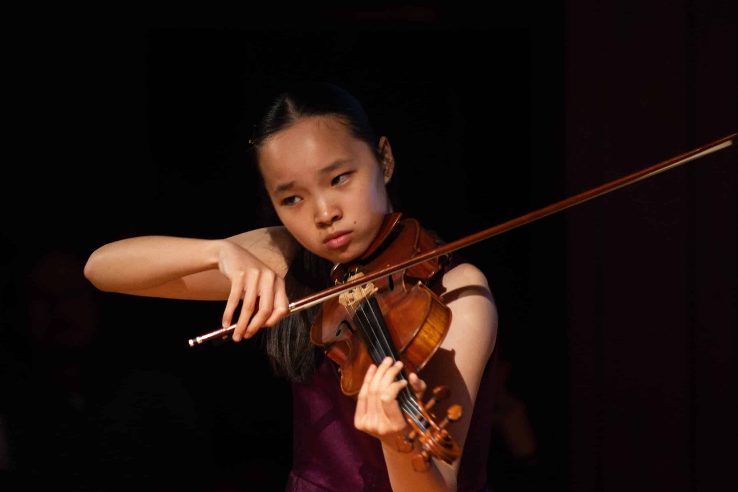 A young woman with long dark hair, wearing a sleeveless top, plays the violin with focus and concentration on a dimly lit stage at the Brentwood International Music Competition.