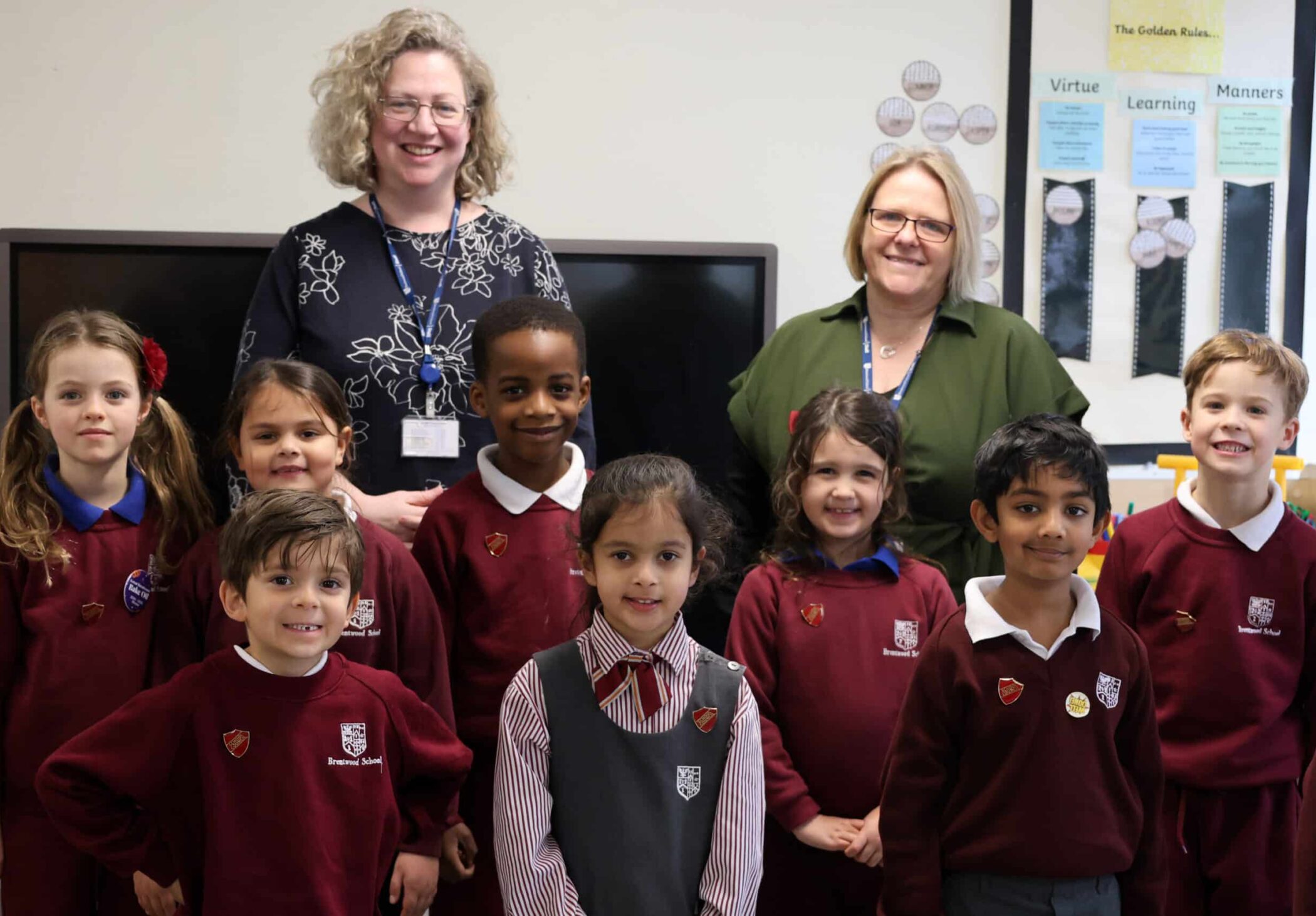 A group of young pupils in maroon uniforms and two adults stand and smile together in a classroom, with posters and rules displayed on the wall behind them.