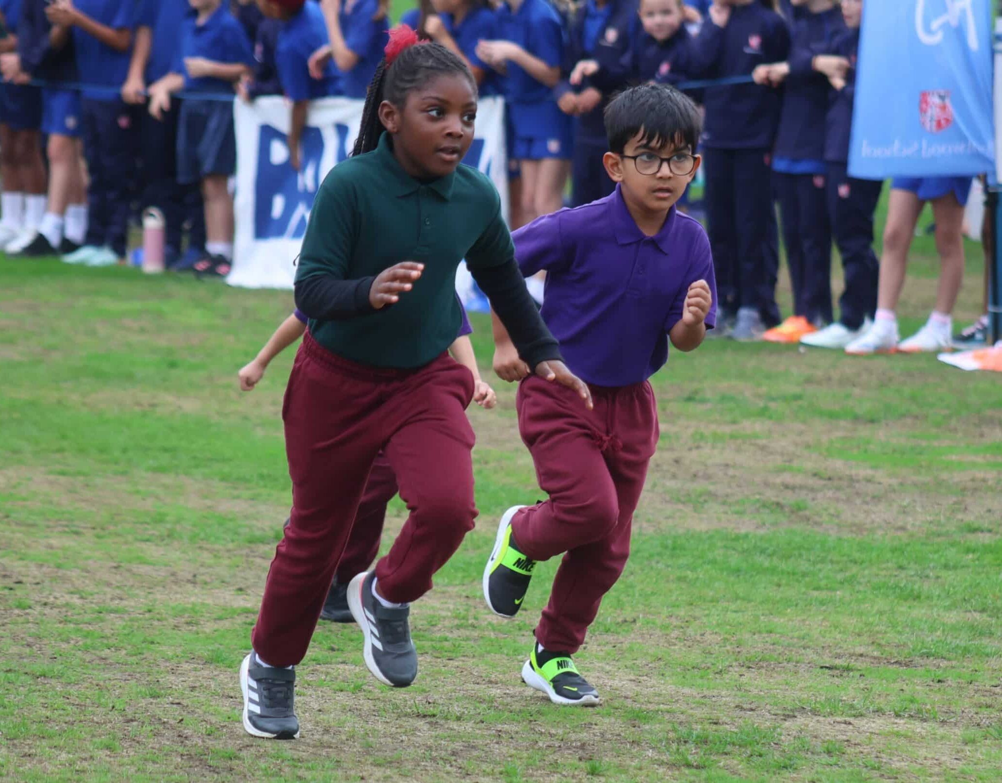 Two children, a girl and a boy, wearing maroon trousers and coloured shirts, run on a grassy field during a race. A crowd of children in blue uniforms watches in the background.