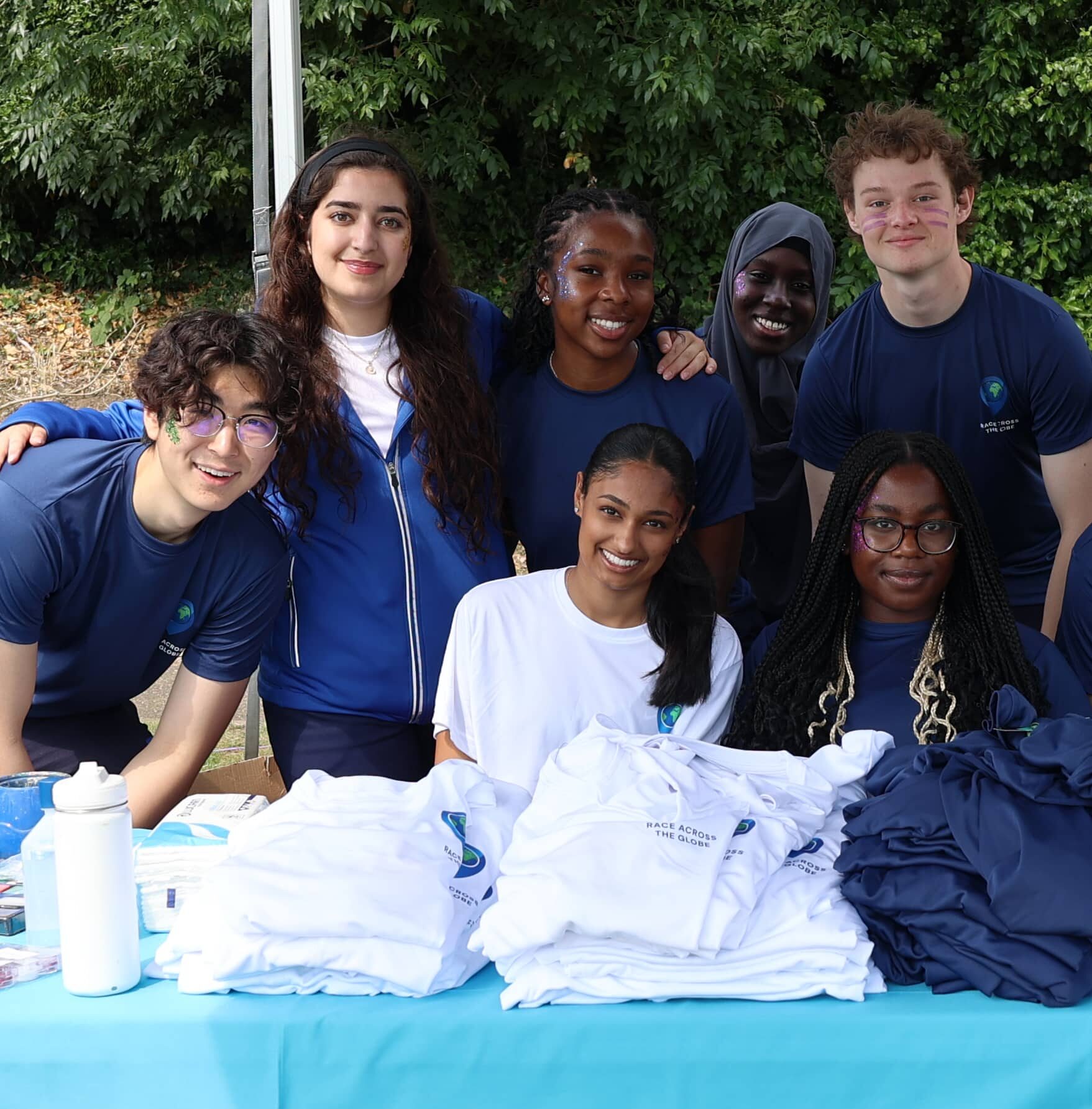 A group of six smiling young adults stand and sit behind a table covered with white boarding school T-shirts and a blue cloth with a red and white crest, outdoors in front of green foliage.