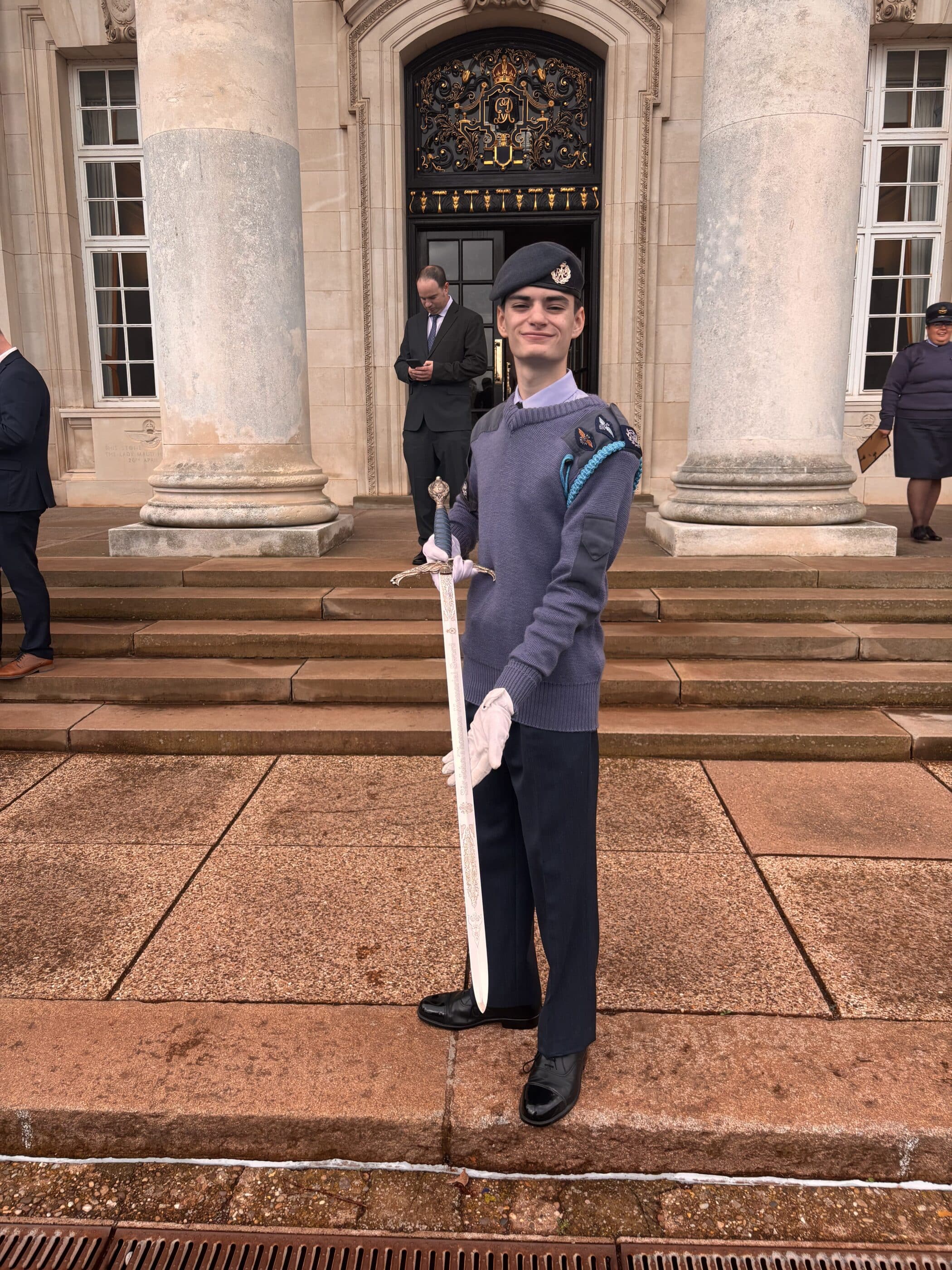 A young cadet in uniform and gloves stands on stone steps in front of a building with columns, proudly holding a ceremonial sword. Two adults are in the background, one reading a mobile, the other standing to the side.