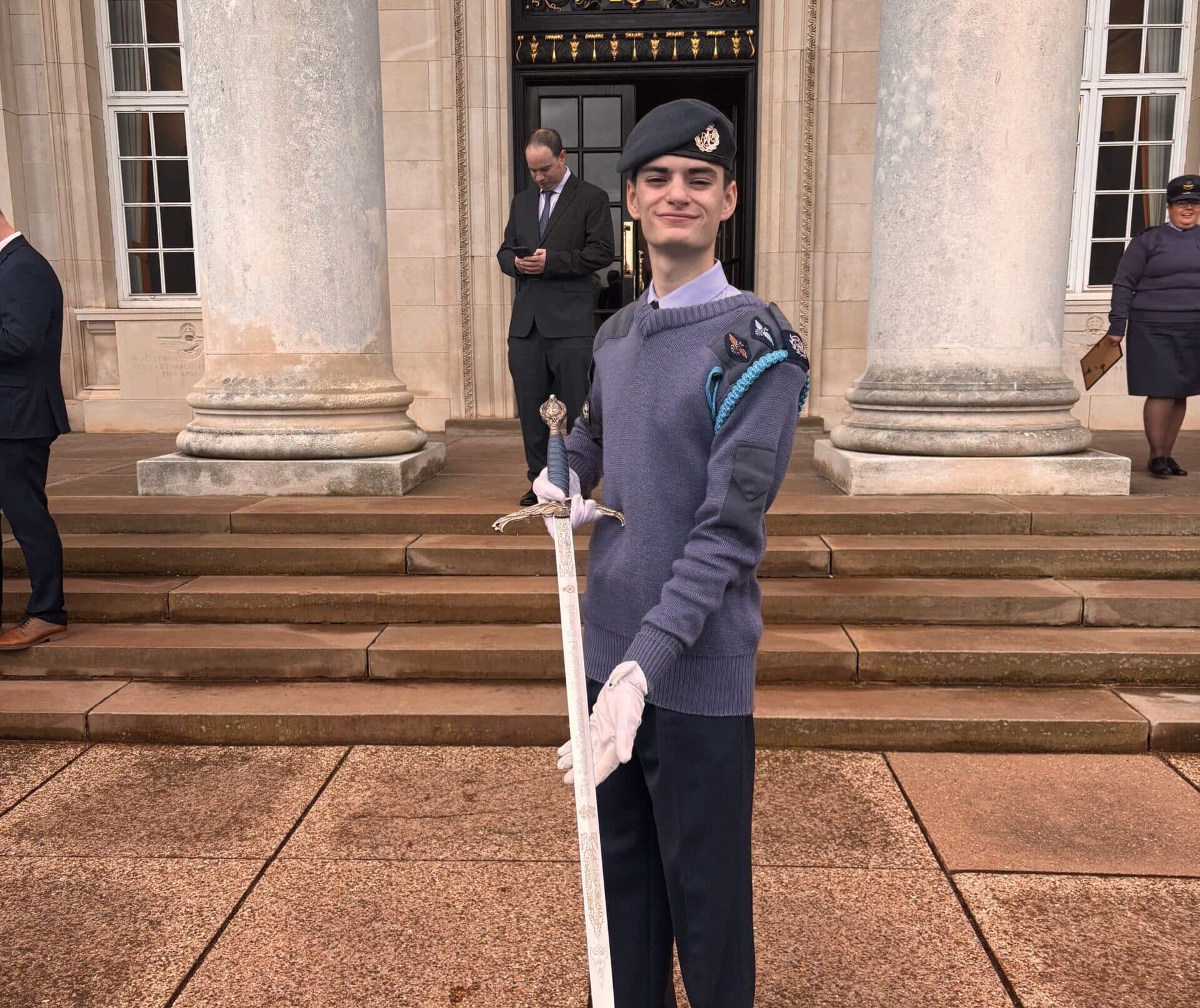 A young cadet in uniform and gloves stands on stone steps in front of a building with columns, proudly holding a ceremonial sword. Two adults are in the background, one reading a mobile, the other standing to the side.