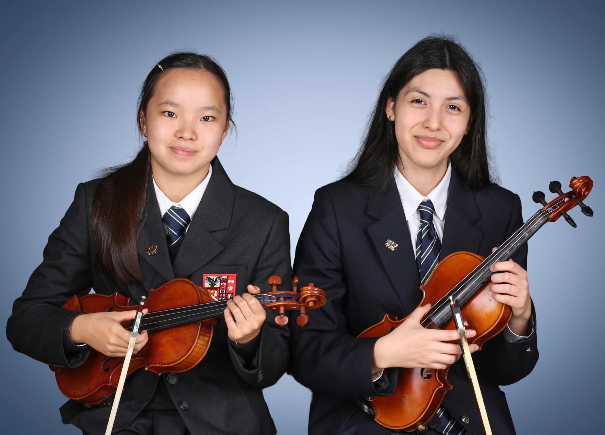 Two students in dark school uniforms, each holding a violin and bow, sit side by side and smile at the camera against a blue-grey background.