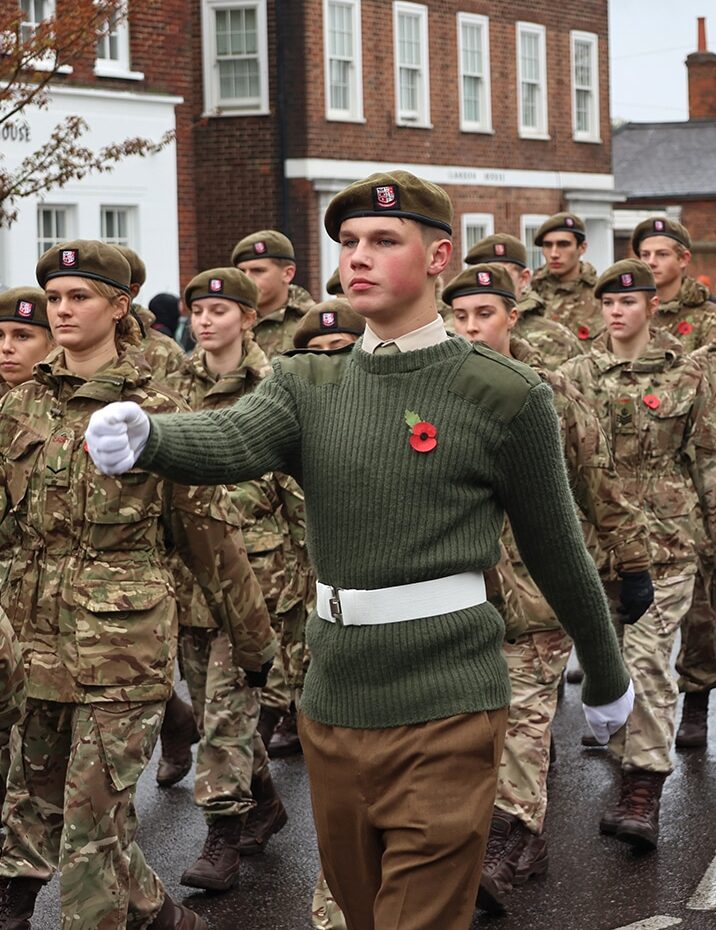A group of young cadets in military uniforms, proudly wearing poppy pins, march in formation as part of a co-curricular activity. The lead cadet in a green jumper and white belt guides them past brick buildings.