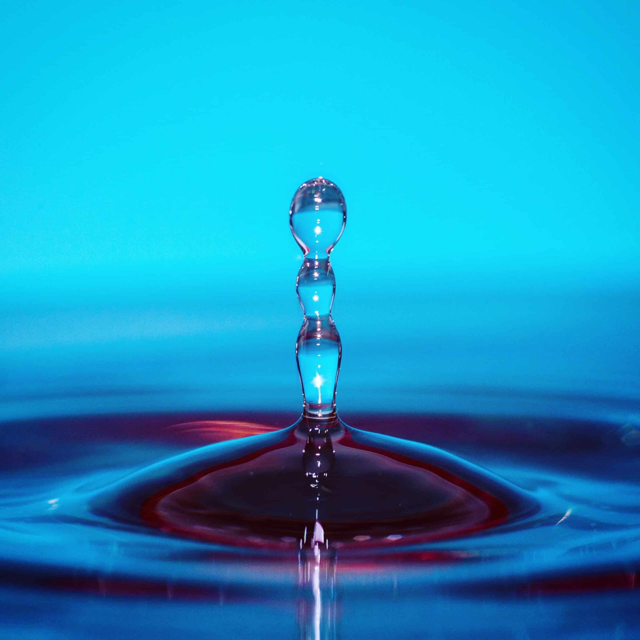 A close-up of a water droplet splashing upward, forming a vertical column with several rounded segments, against a bright blue background. Ripples spread across the surface below the droplet.