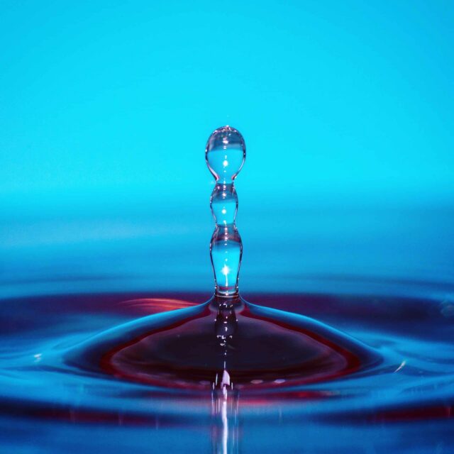 A close-up of a water droplet splashing upward, forming a vertical column with several rounded segments, against a bright blue background. Ripples spread across the surface below the droplet.