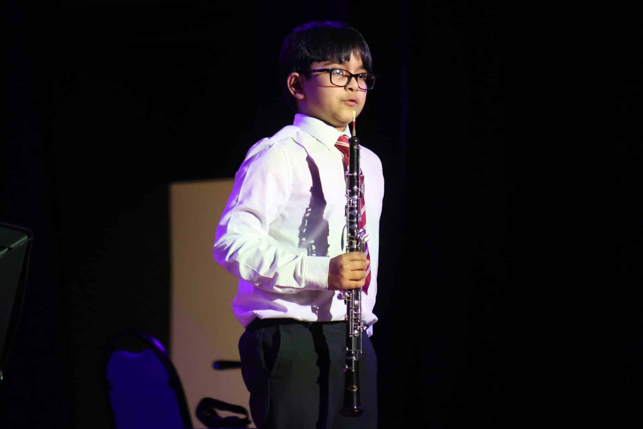 A young boy wearing glasses, a white shirt, and a red-striped tie stands on stage holding an oboe, with a chair and dark background behind him.