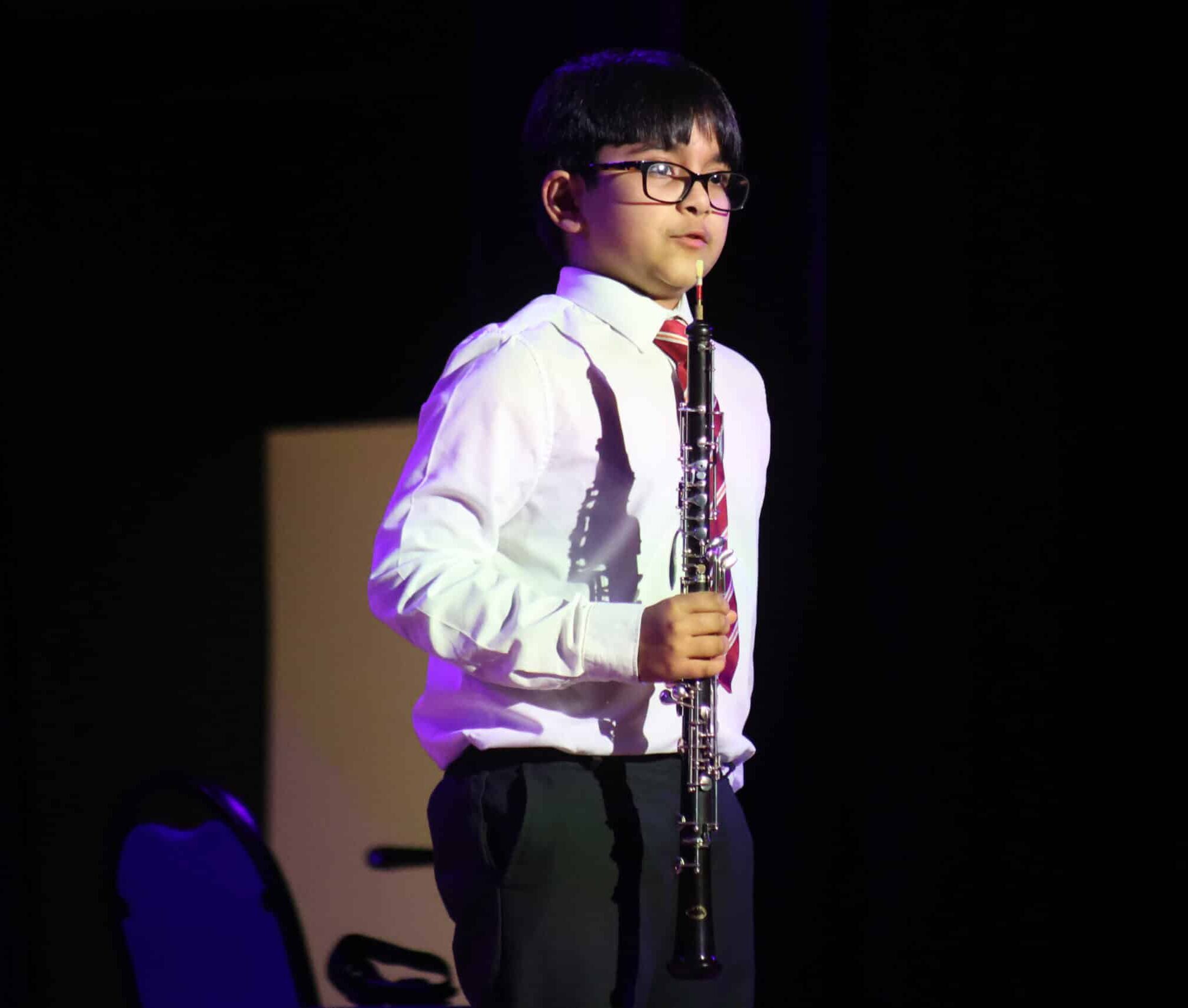A young boy wearing glasses, a white shirt, and a red-striped tie stands on stage holding an oboe, with a chair and dark background behind him.