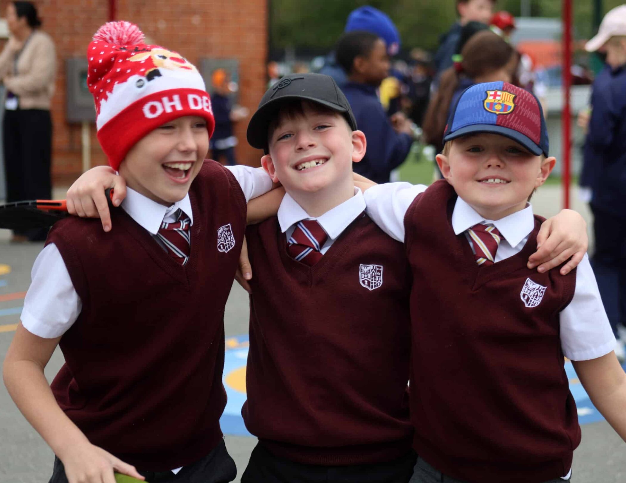 Three boys in school uniforms, wearing maroon jumpers and ties, stand arm in arm smiling. One wears a red hat, another a black cap, and the third a blue and red cap. They are outdoors with other children in the background.