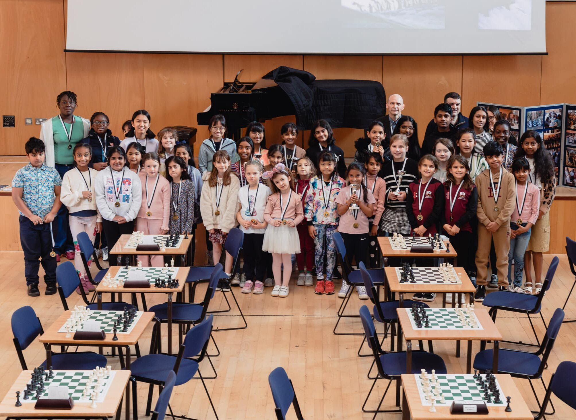 A large group of children and a few adults pose for a photo in a hall with chessboards set up on tables. Many children wear medals, suggesting a chess tournament or competition. A piano and posters are in the background.