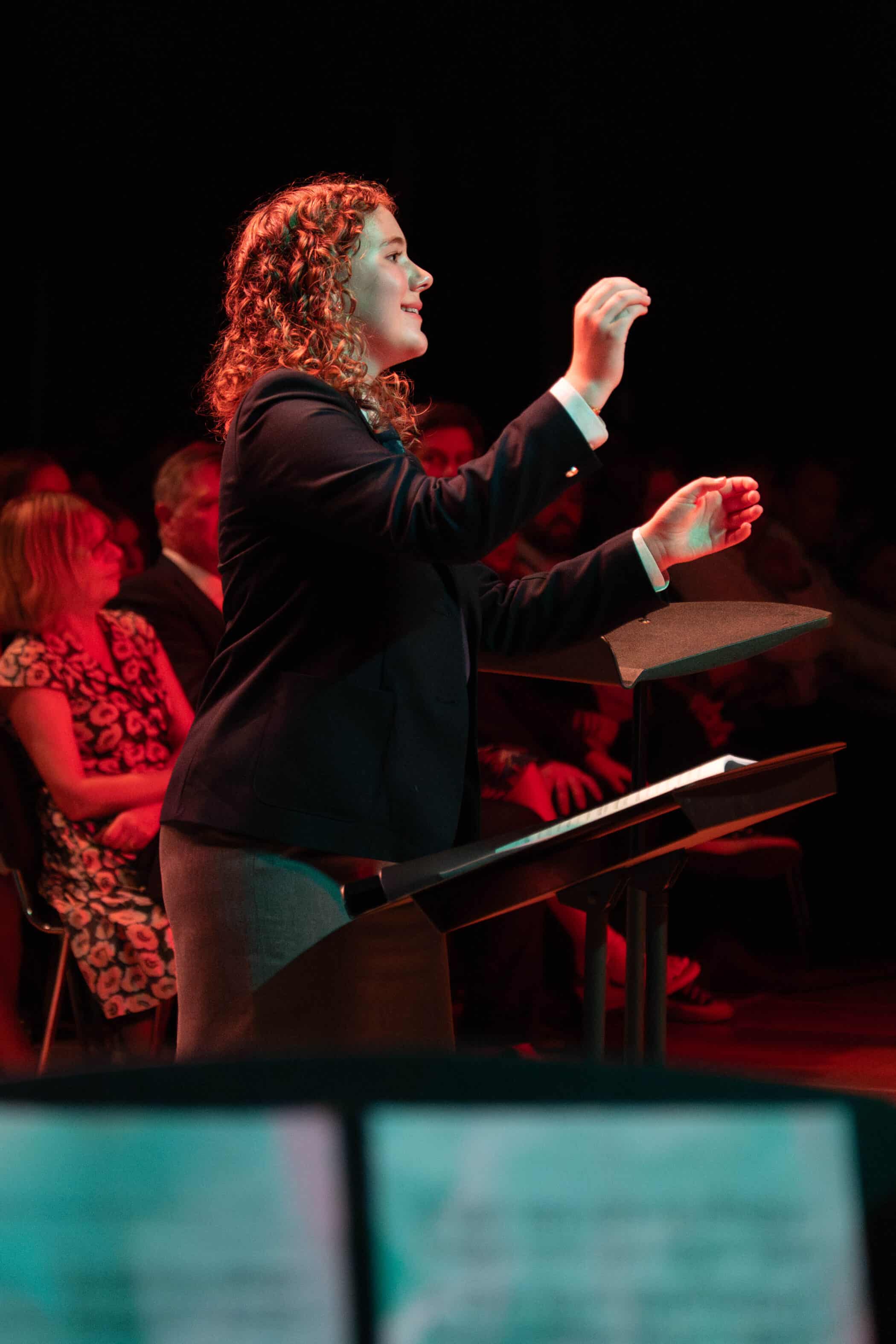 A woman with curly hair in a suit conducts music, facing an audience in a dimly lit room with a red hue. She gestures with both hands, appearing focused and engaged. People sit in the background watching.