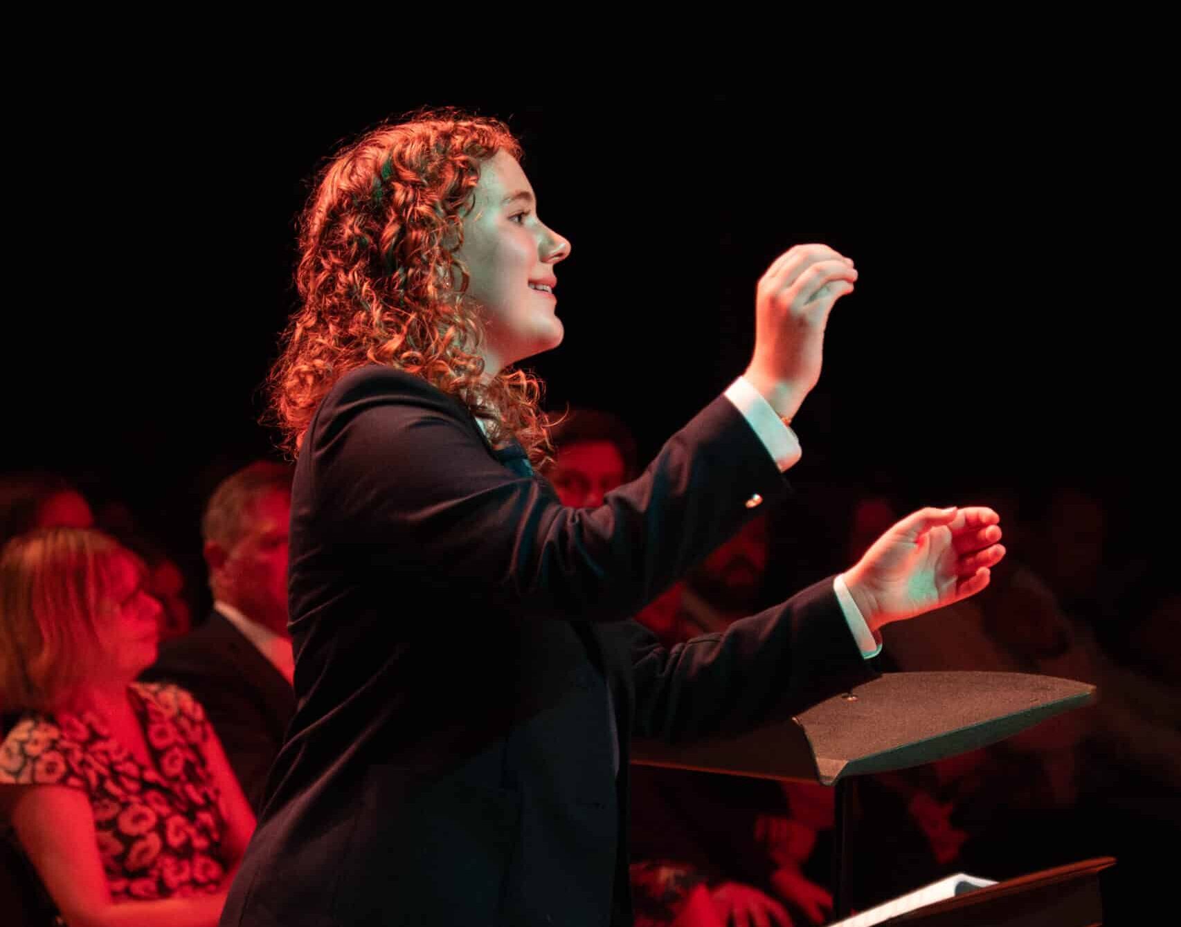 A woman with curly hair in a suit conducts music, facing an audience in a dimly lit room with a red hue. She gestures with both hands, appearing focused and engaged. People sit in the background watching.