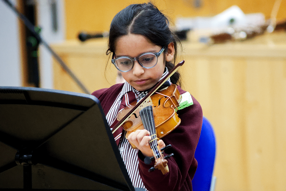 A young girl with glasses and dark hair plays the violin, looking intently at sheet music on a stand. She wears a striped shirt and maroon cardigan, seated indoors with a blurred background.