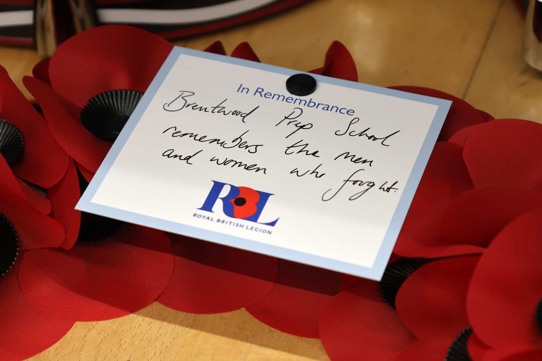 A handwritten note reading, Brentwood Prep School remembers the men and women who fought, is clipped to a wreath of red poppies. The note includes the Royal British Legion logo and the heading In Remembrance.