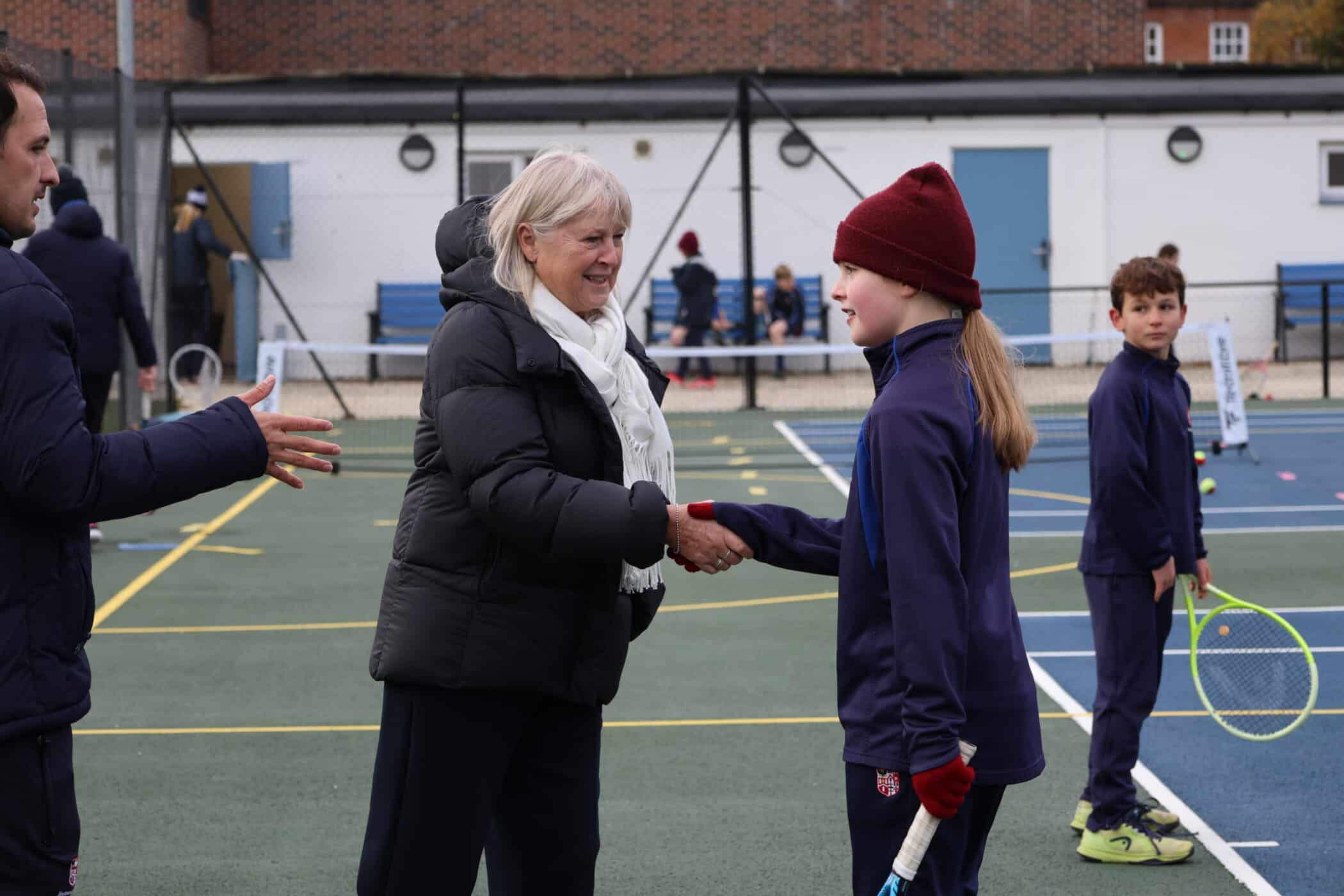 An older woman in a black coat and white scarf shakes hands with a young girl in a red beanie and navy sports kit on an outdoor tennis court. Other people sit on benches in the background.