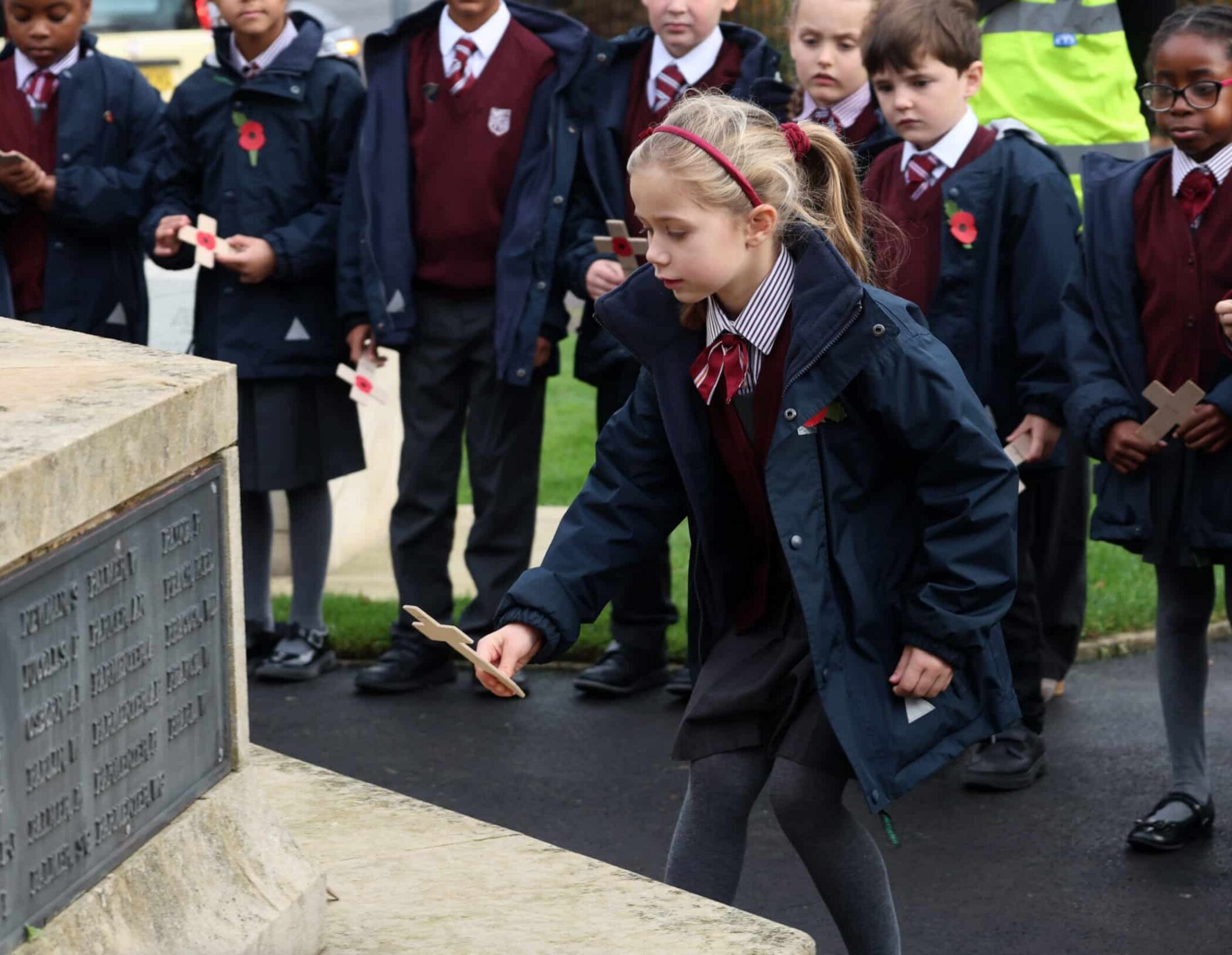 A young girl in school uniform places a wooden cross at a stone memorial, whilst other schoolchildren in similar uniforms stand behind her, observing the ceremony.