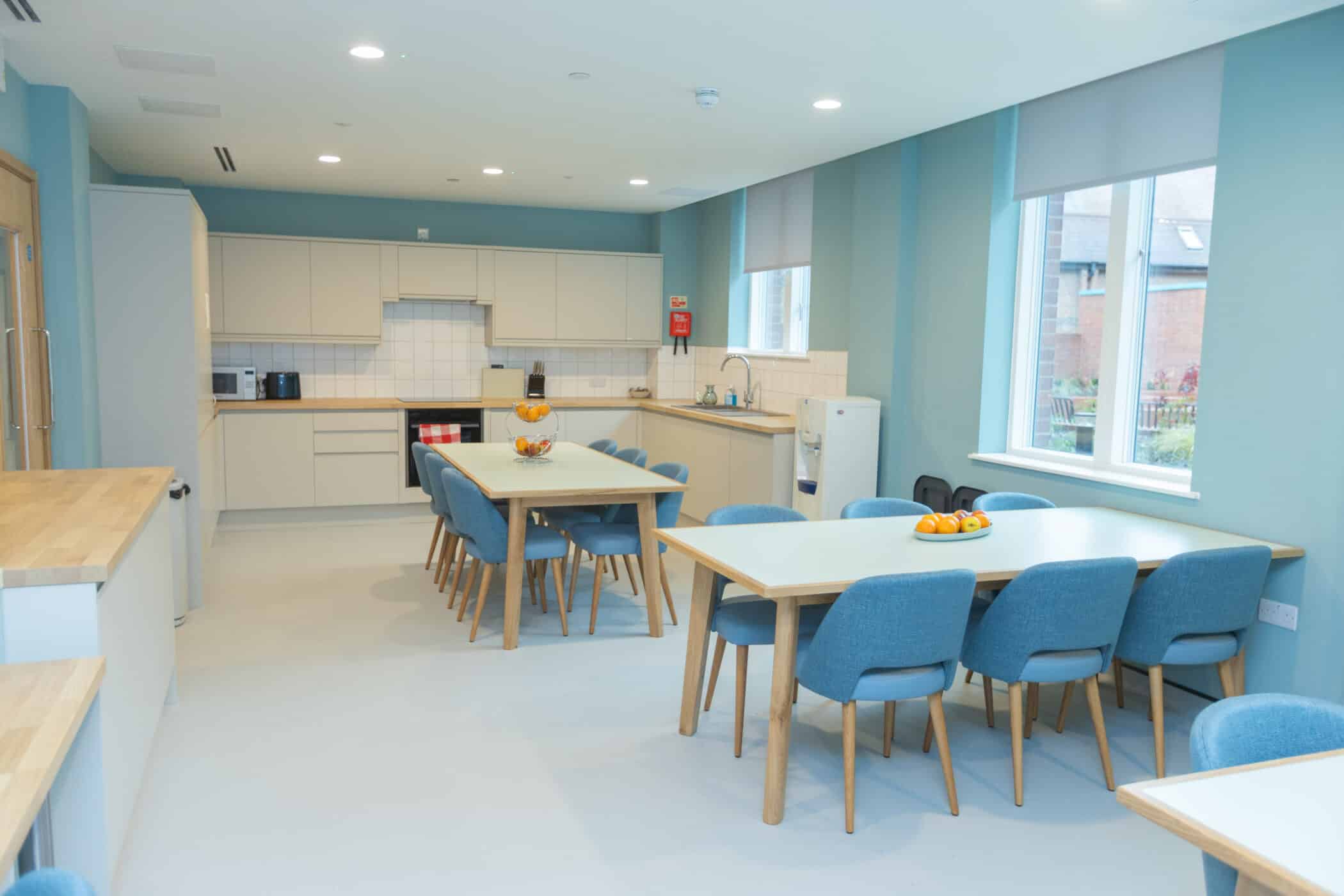 Modern communal kitchen in the New Boarding House features light-coloured cupboards, two rectangular tables with grey chairs, a bowl on one table, tiled splashback, and large windows that fill the space with natural light.