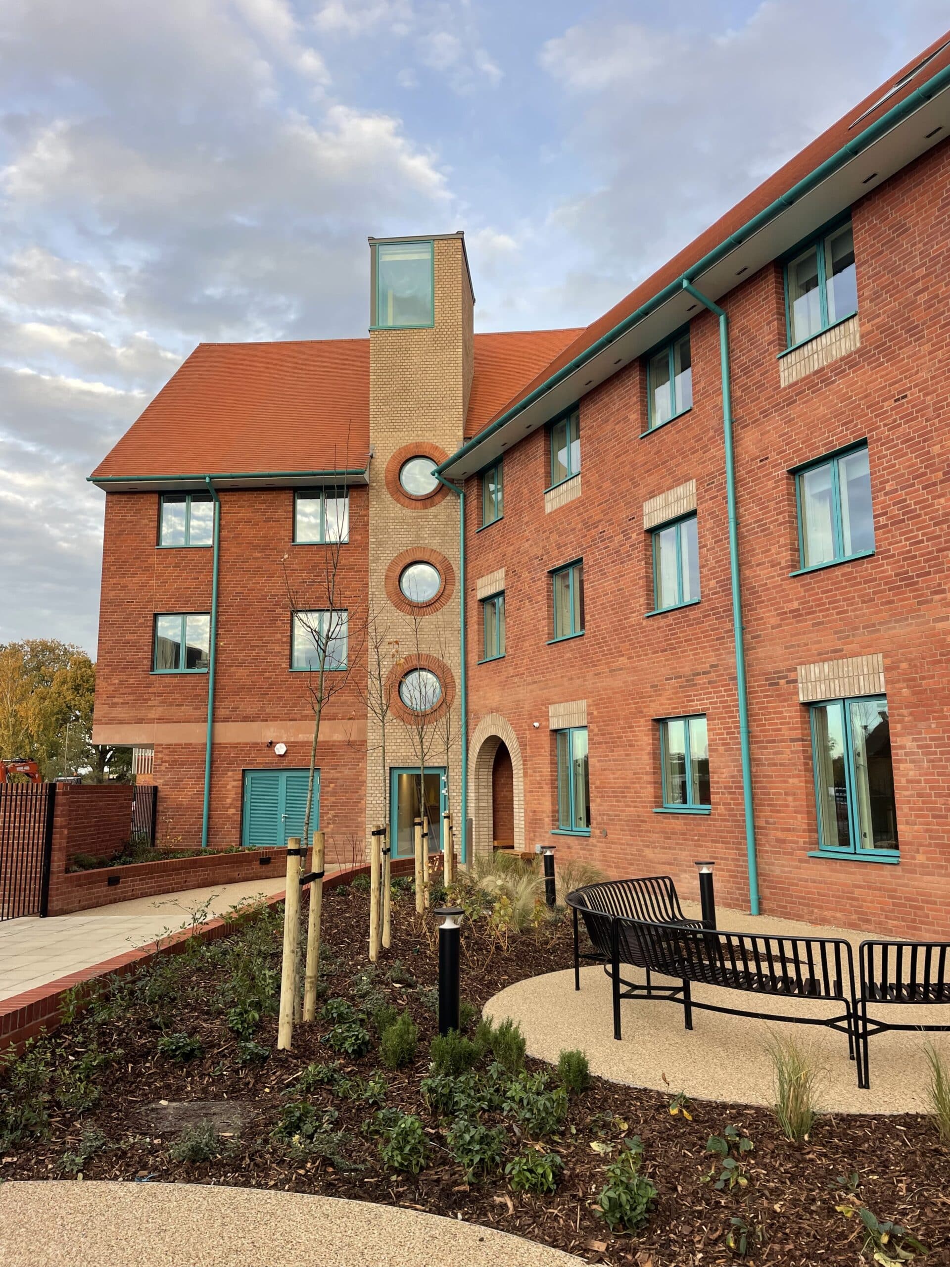 A modern red-brick building with teal window frames and circular windows, surrounded by landscaped gardens -perfect for those experiencing boarding at Brentwood -with a bench, young trees, and a curved pathway under a partly cloudy sky.