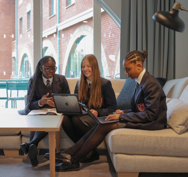 Three students in school uniforms sit on a sofa in a modern room at Brentwood, working together on laptops and notes. Smiling and engaged in discussion, they embody the collaborative spirit of boarding at Brentwood. Large windows and a lamp are behind them.