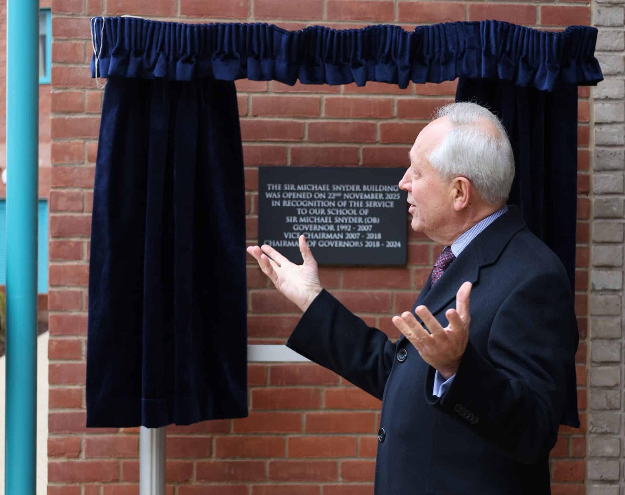 An older man in a suit gestures with open hands while standing in front of a plaque on a brick wall, partially framed by blue curtains above and beside the plaque.