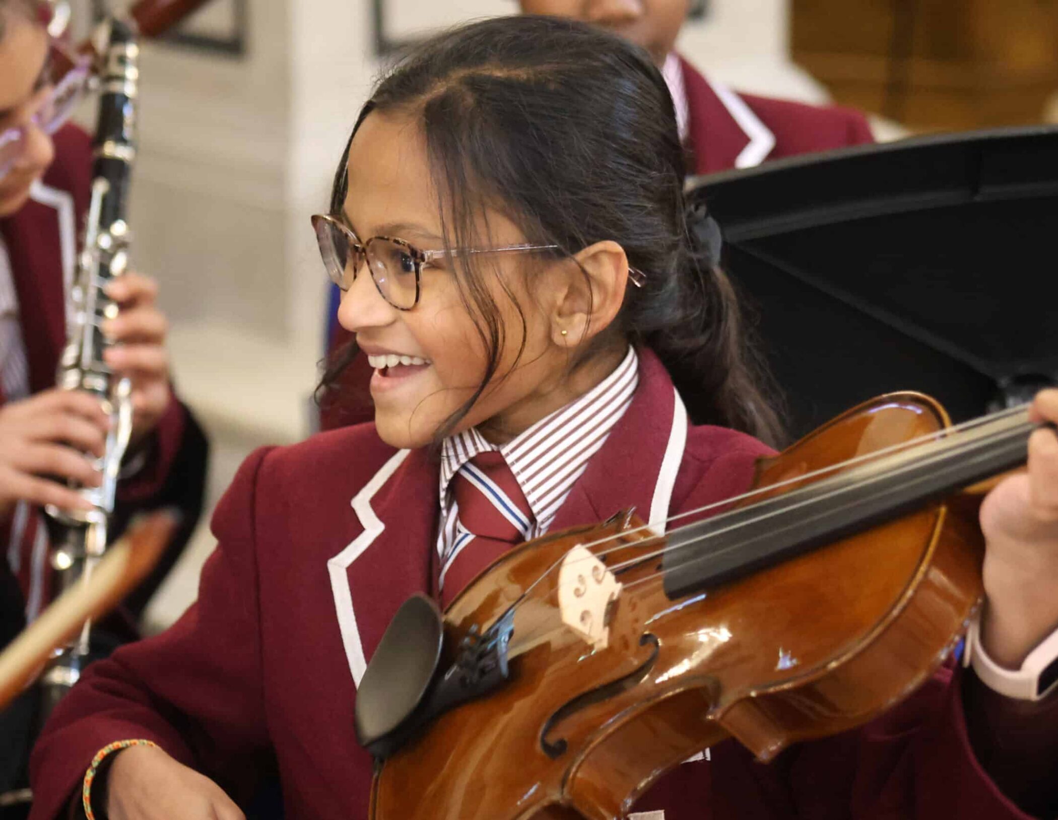 A smiling student in a maroon blazer and glasses holds a violin, appearing to play with an orchestra. Other young musicians are visible in the background, dressed in matching uniforms.