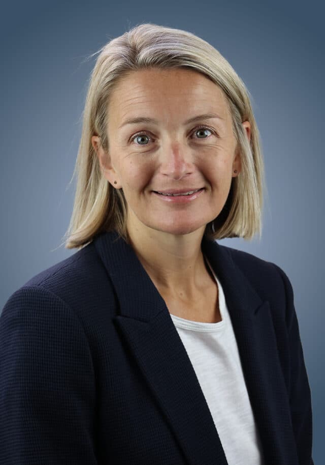 A woman with straight blonde hair wearing a navy blazer over a white top is smiling softly against a plain blue background.