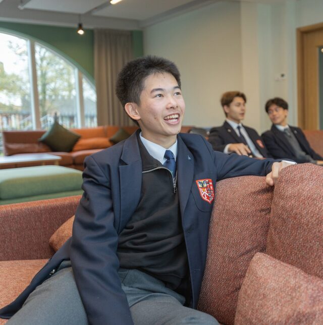 A smiling student in a school blazer sits on a sofa in a lounge area, reflecting the warm atmosphere of Boarding at Brentwood, with two other students chatting nearby. Large windows and modern furniture create an inviting space.