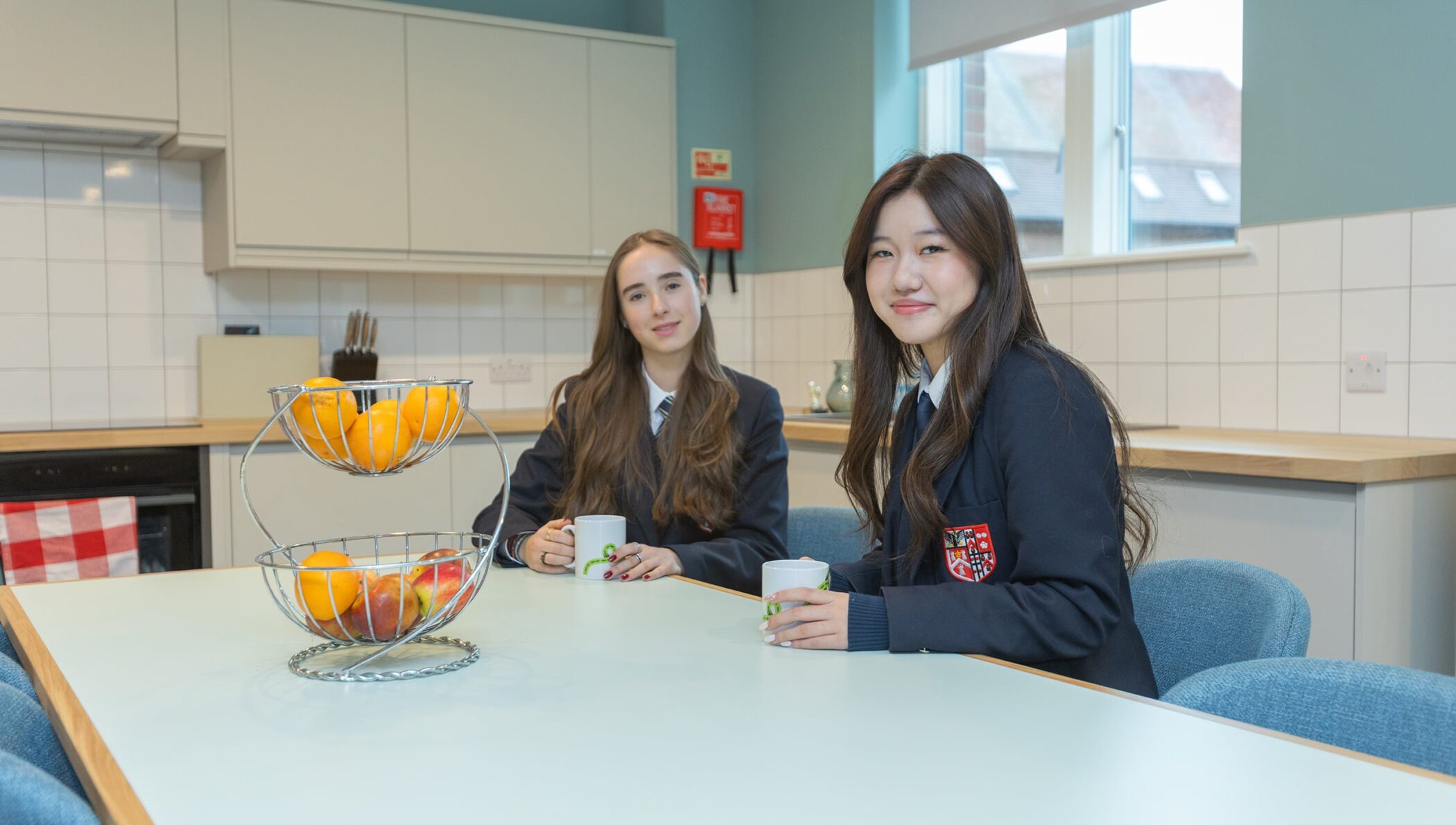 Two young boarders in Brentwood School uniforms sit at a kitchen table with mugs in hand. A fruit stall brimming with oranges and apples sits between them, set against light blue walls and cupboards.