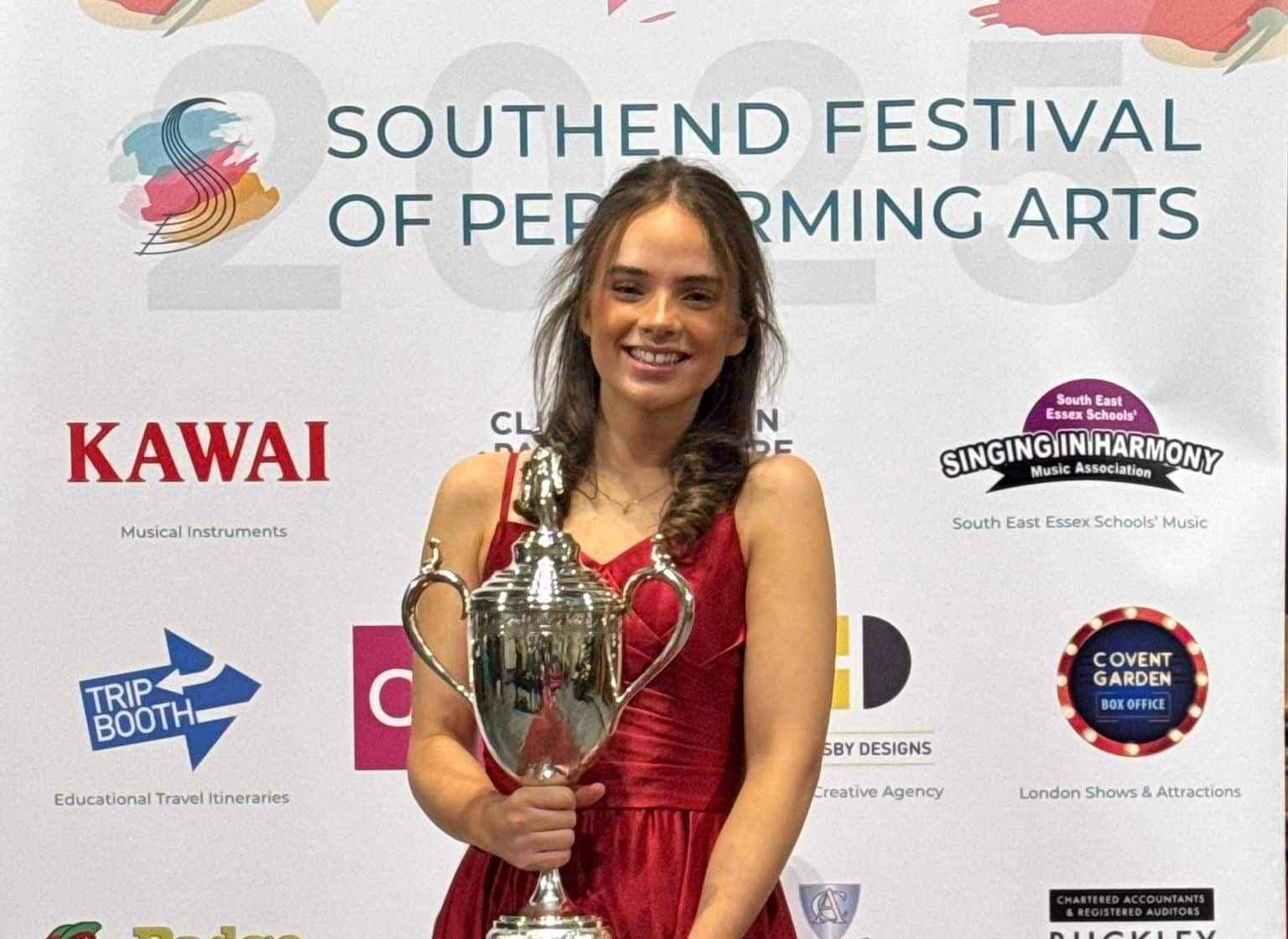 A young woman in a red dress smiles whilst holding a large silver trophy in front of a Southend Festival of Performing Arts banner featuring various sponsor logos.