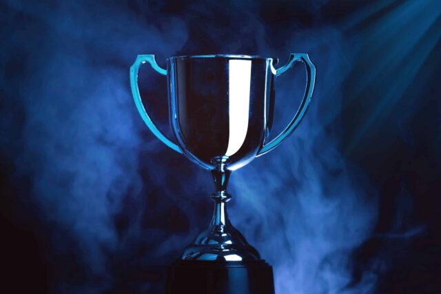 A shiny silver trophy with two handles is centred against a dramatic blue and smoky background, illuminated by a beam of light from the upper right corner.