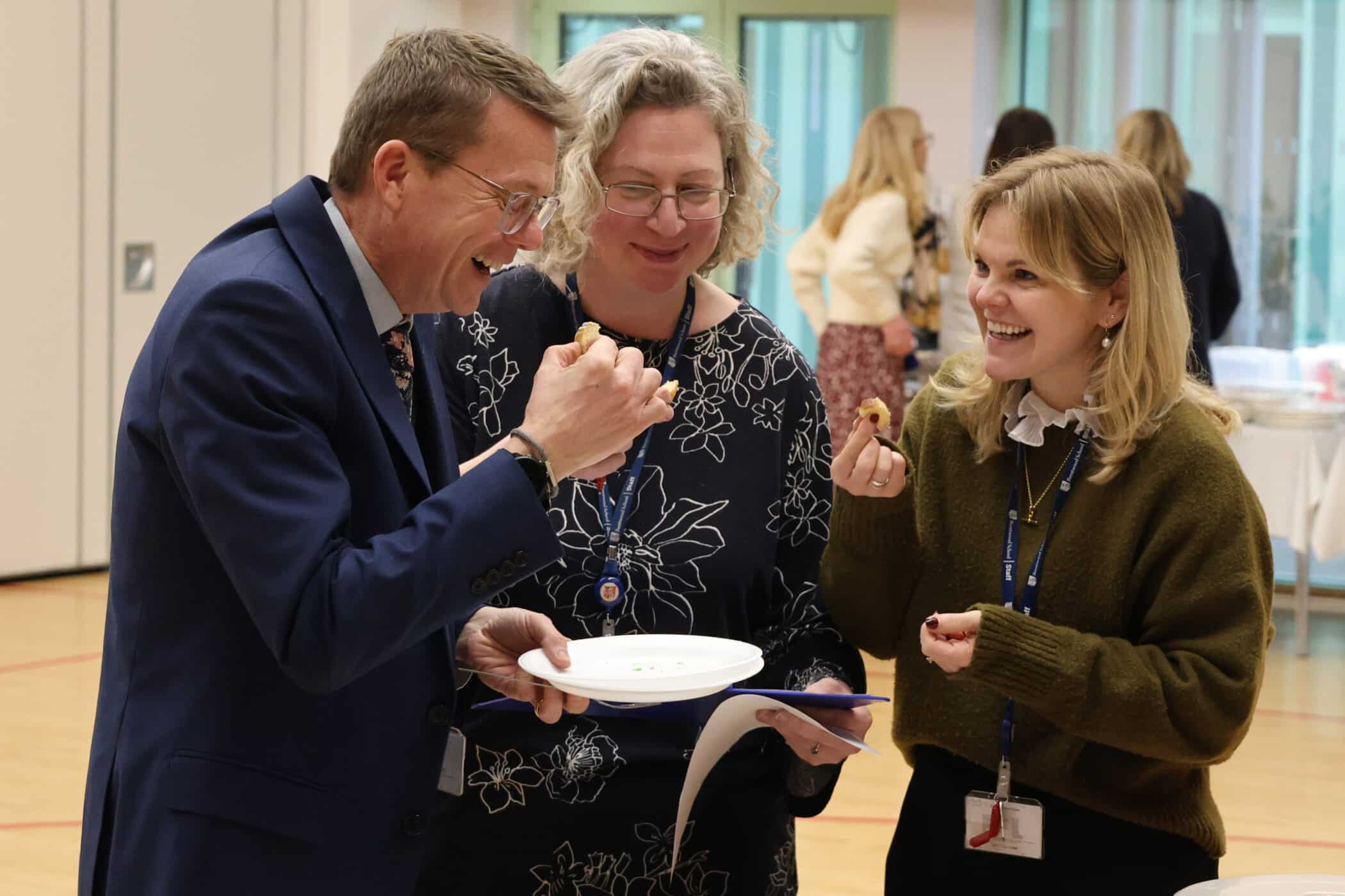 Three adults stand together indoors, smiling and laughing whilst holding food on paper plates. Two women and one man appear to be enjoying a light-hearted moment at a social or professional gathering.