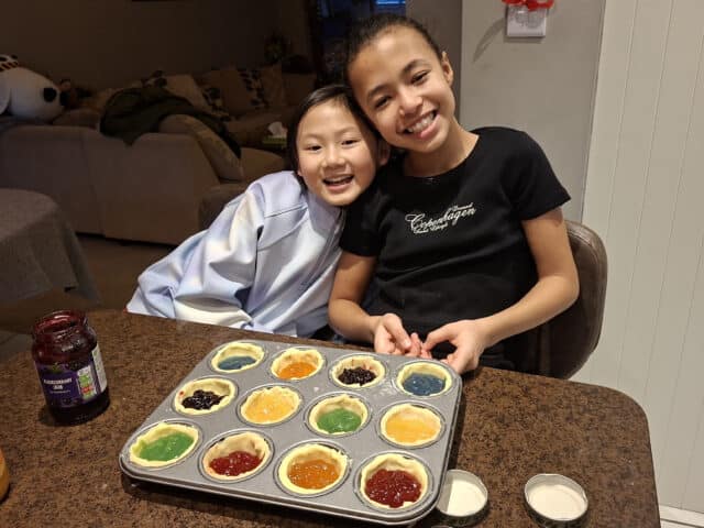 Two smiling children sit at a kitchen worktop, holding a muffin tin filled with colourful unbaked tarts of various jams and fillings. A jar of jam and two empty bowls are on the brown work surface.