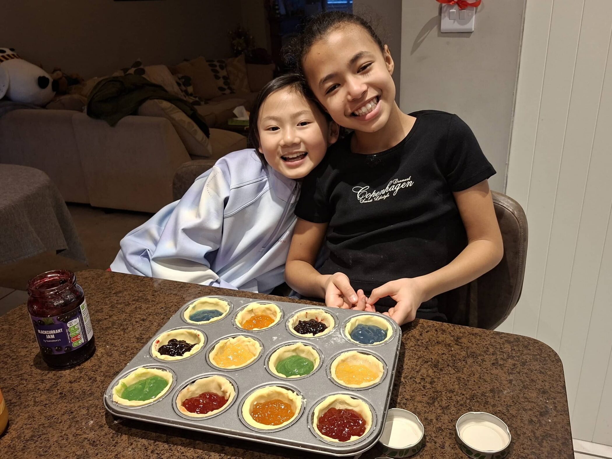 Two smiling children sit at a kitchen worktop, holding a muffin tin filled with colourful unbaked tarts of various jams and fillings. A jar of jam and two empty bowls are on the brown work surface.