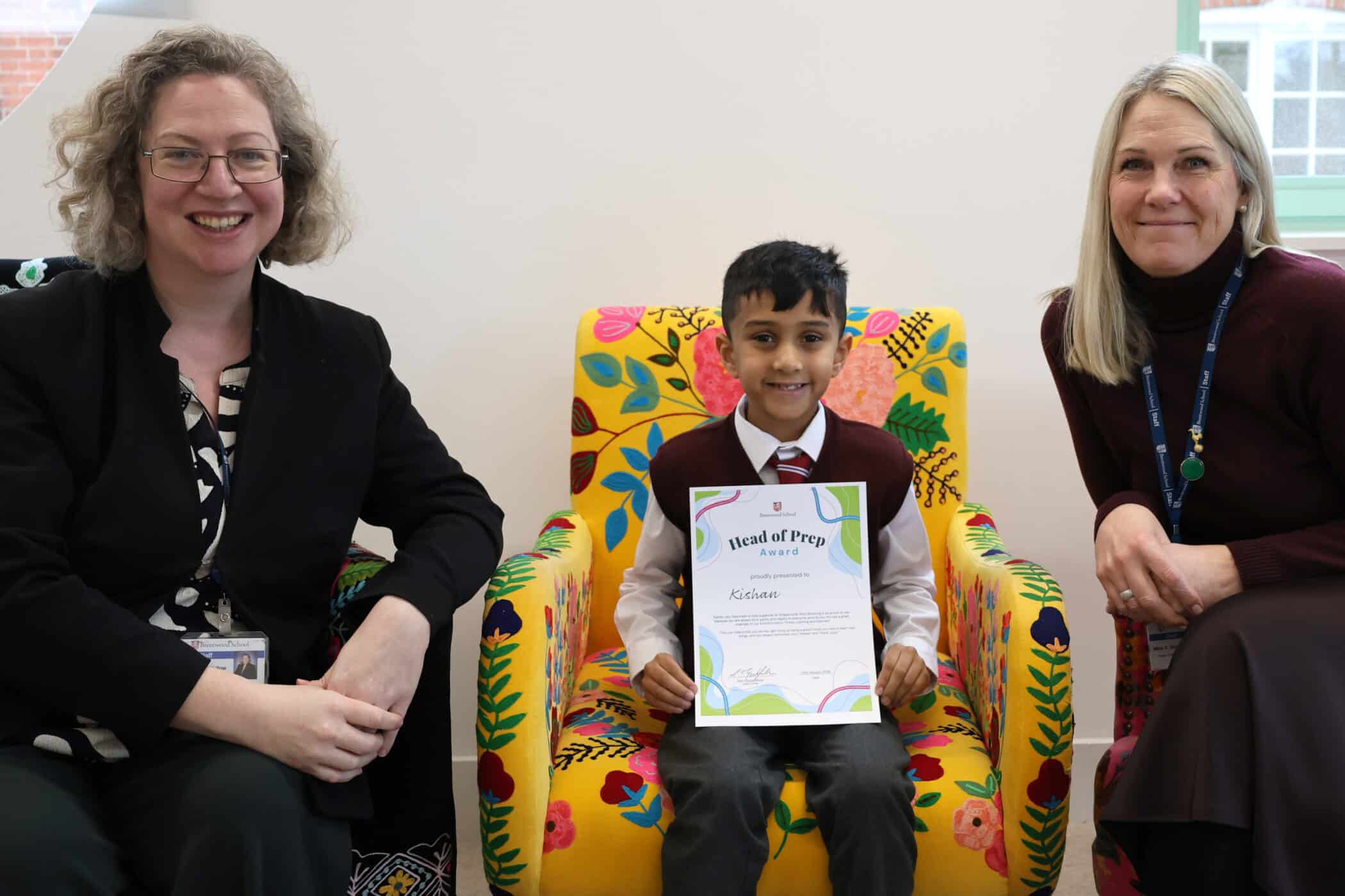 A young boy sits on a colourful floral chair, smiling and holding a certificate, with two women sitting beside him, also smiling, in a bright indoor setting.