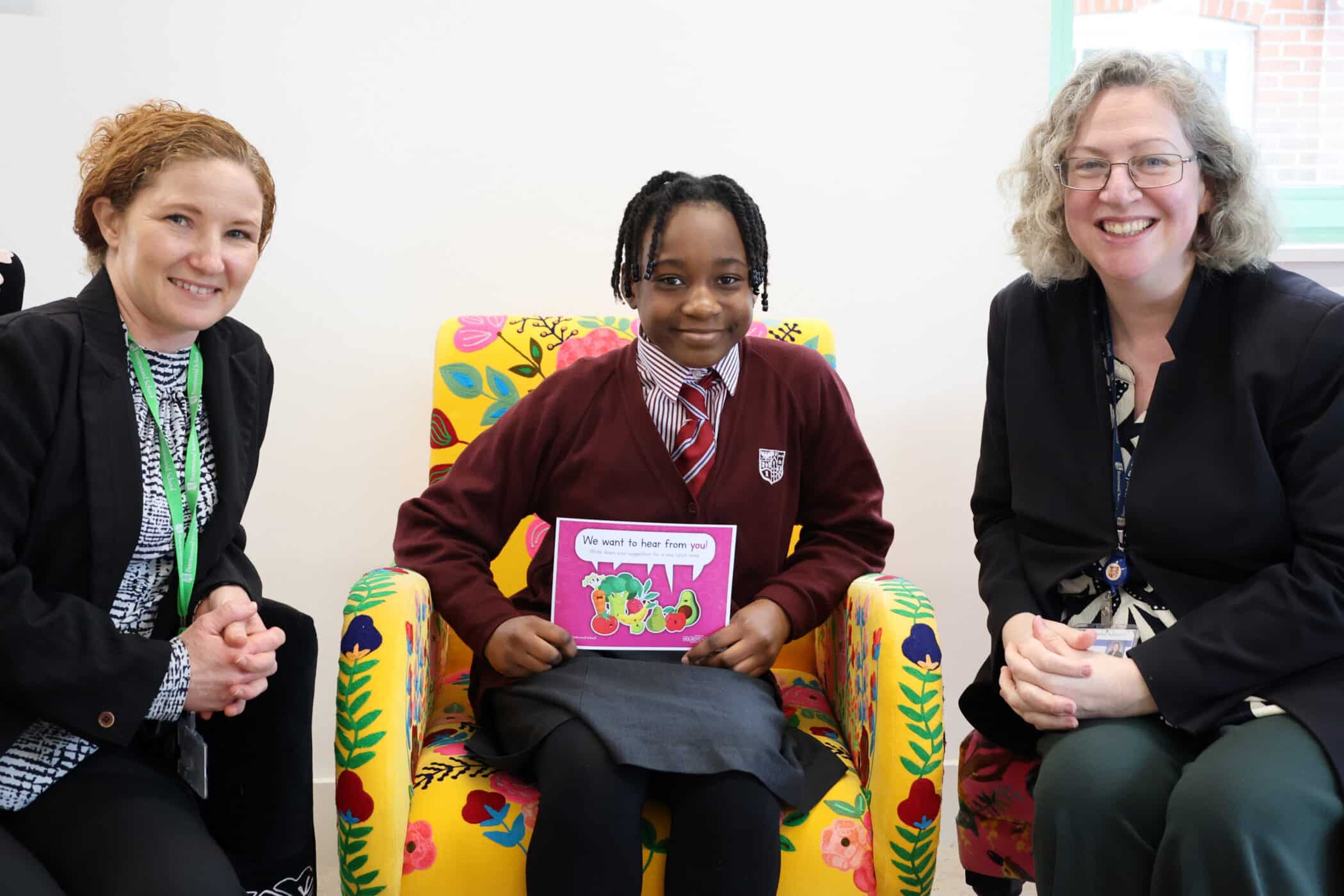 Three people sit together, smiling. A girl in a school uniform sits on a colourful chair, holding a sign that says, We want to hear from you. Two women, one on either side, are dressed in business attire.