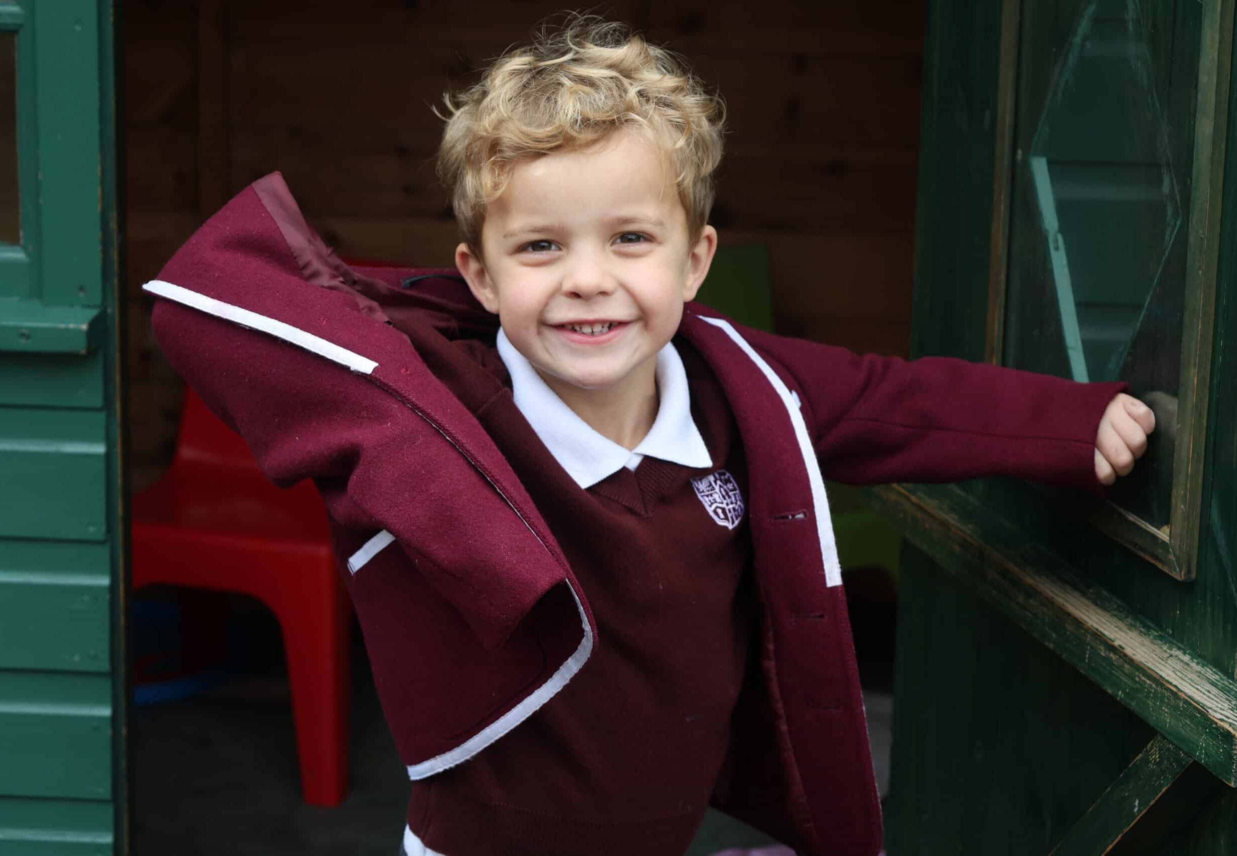 A young child with curly blonde hair smiles while putting on a maroon school blazer over a white polo shirt, standing in a playhouse doorway.