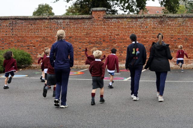 A group of young children in maroon school uniforms and three adults walk and play on a playground near a brick wall, some children running ahead and others walking together.