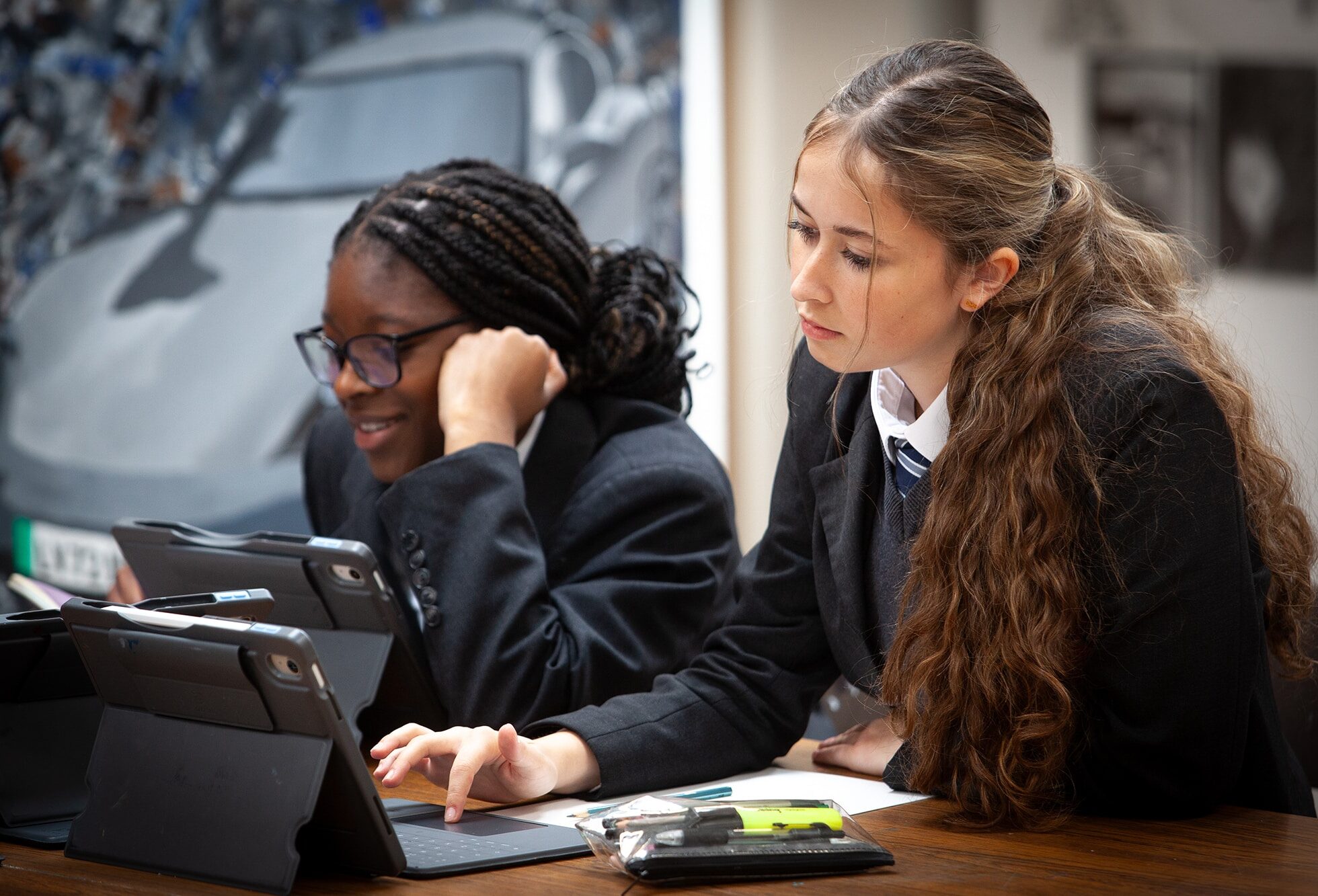 Two pupils in school uniforms work on tablets at a desk. One, with plaited hair and glasses, smiles, while the other, with long wavy hair, focuses intently on the screen during their academic activities. School supplies are on the table.