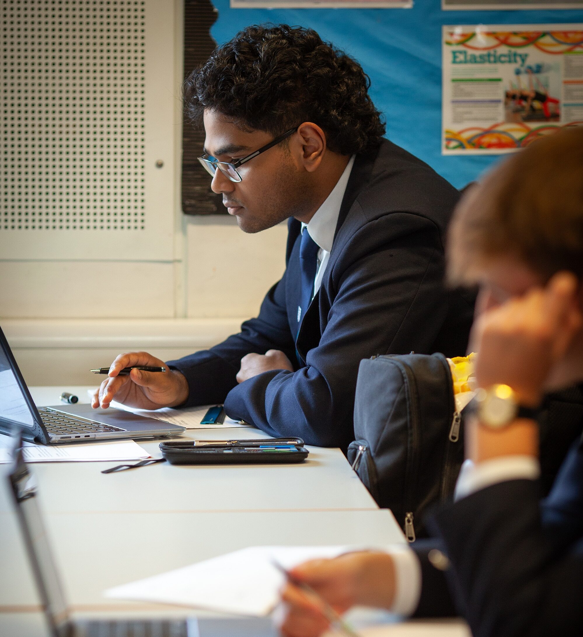 A student in a suit sits at a desk using a laptop, pen in hand and focused on his work, an ideal example of dedication to Sixth Form. Another student writes notes in the blurred foreground, with classroom posters visible behind them.