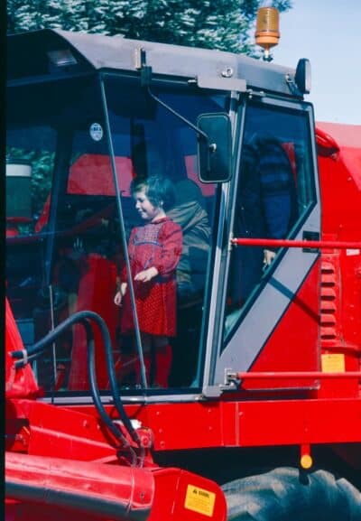 A young child in a red dress stands inside the cab of a large red tractor or combine harvester, looking out through the glass, with an adult partially visible behind her. Trees and blue sky are in the background.