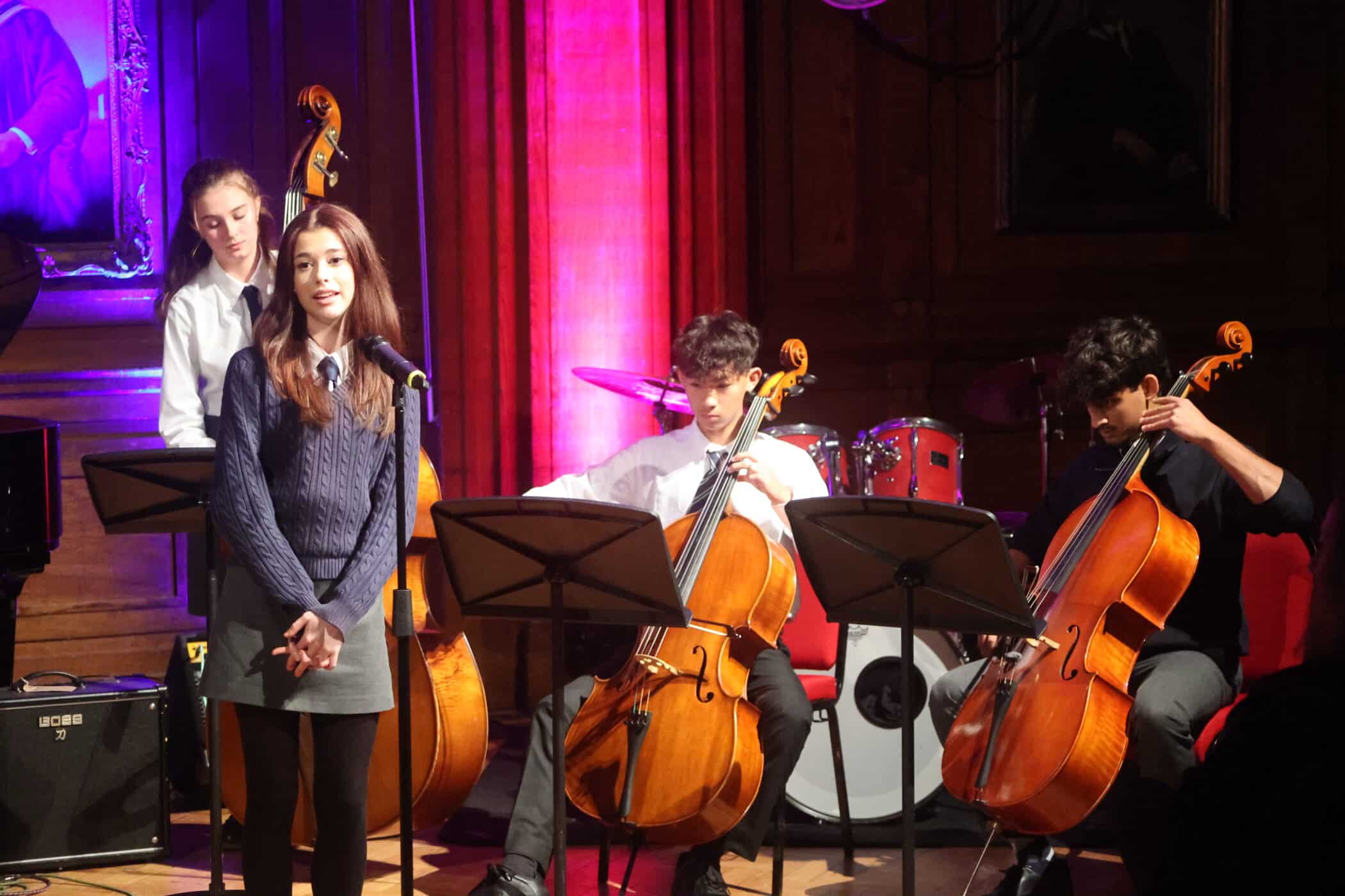 A young woman stands at a microphone, smiling, whilst three musicians behind her play cellos on stage at Brentwood School. The background features red curtains, stage lights, and a drum kit.